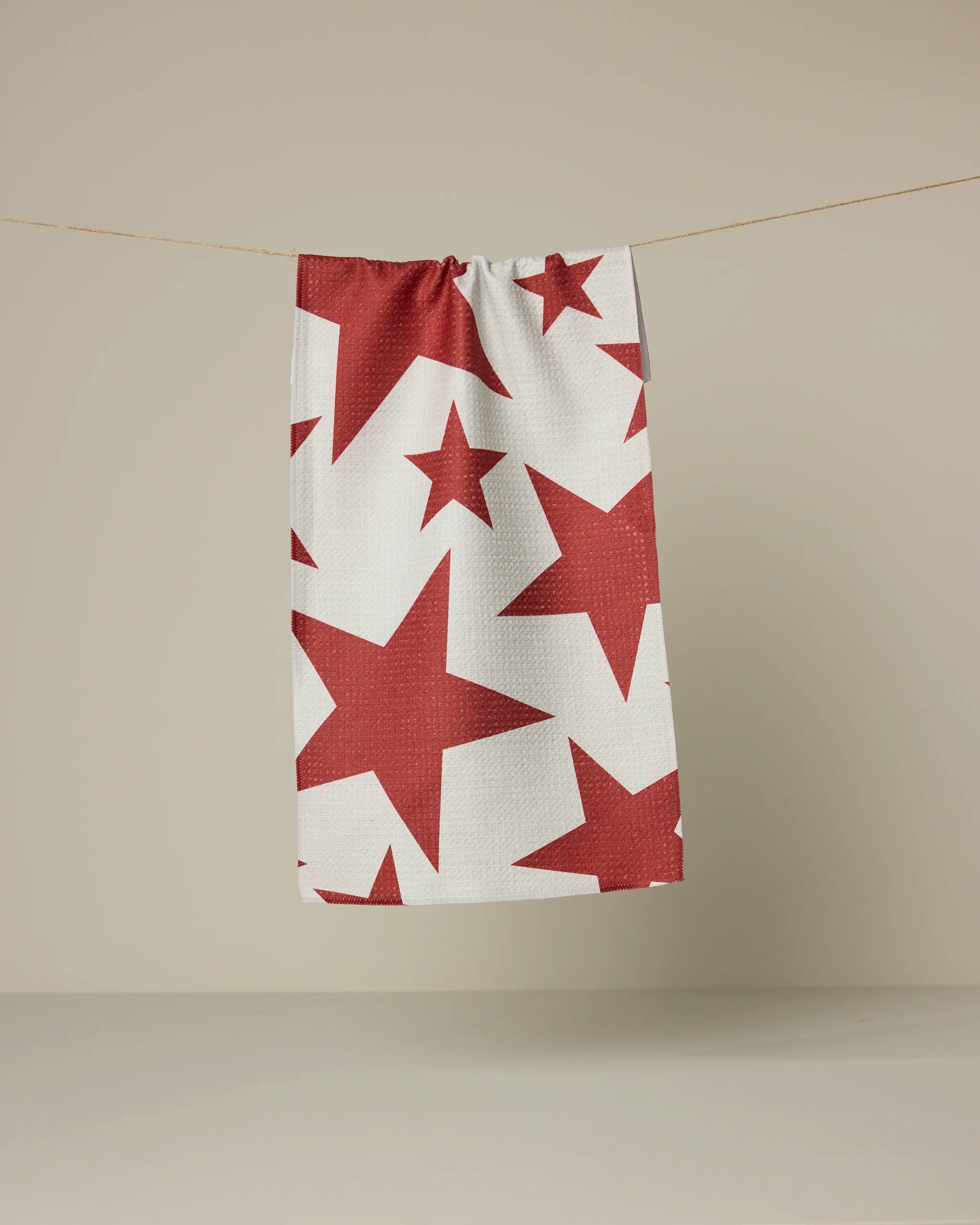 The Geometry Celestial towel, ultra-absorbent and white with red stars in various sizes, hangs on a clothesline against a plain beige background.