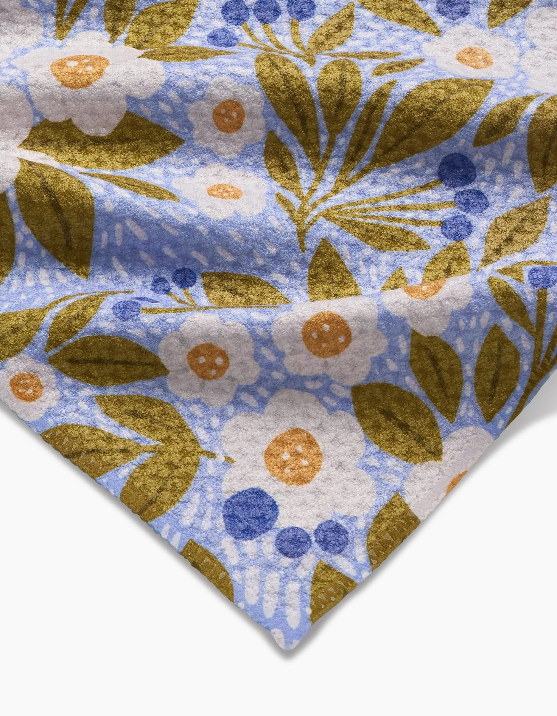 A close-up of the Lisa Nelson Chamomile And Blue Bar Towel made from Geoweave fabric, showcasing a floral print with white flowers, golden yellow leaves, orange centers, and blue accents on a light blue background. The corner is folded.