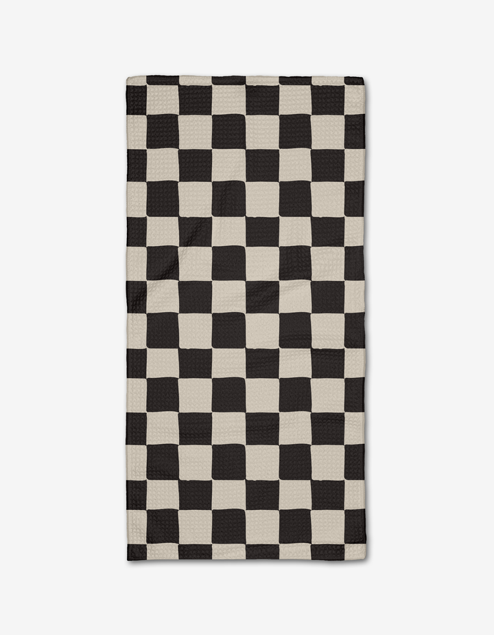 The Checkmate Bar Towel by Danelys Sidron, made of ultra-absorbent Geoweave fabric, showcases a black and off-white wavy checkerboard design that creates an optical illusion and is pictured flat on a white background.