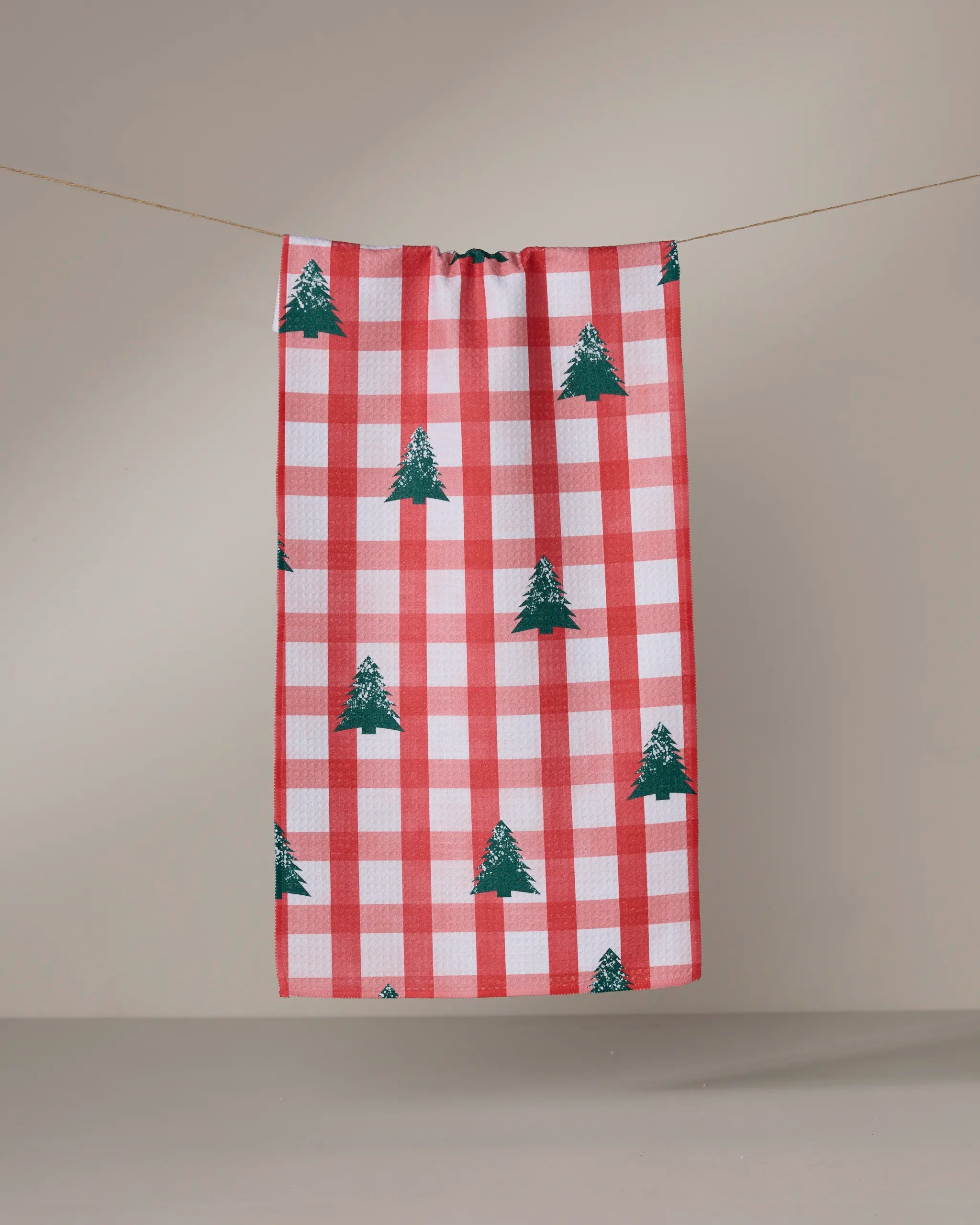 The Geometry "Christmas Pines" towel features red and white checks with green Christmas tree patterns, hanging on a clothesline against a light background.