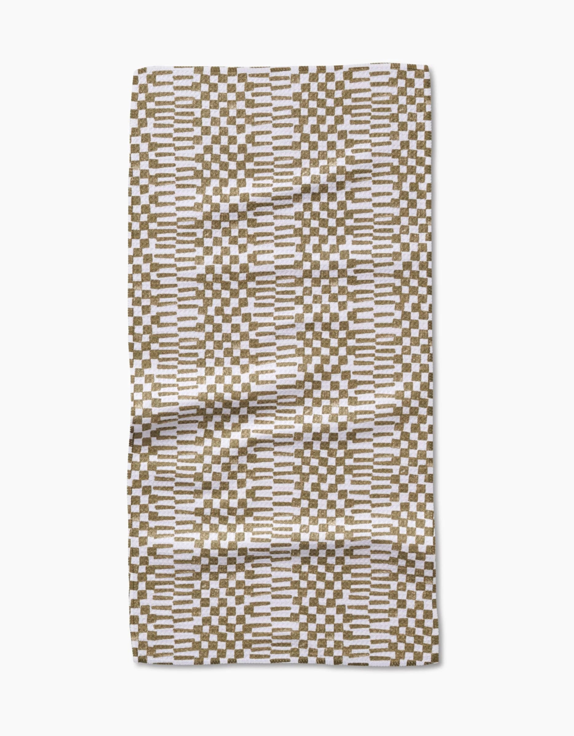 The Studio Jeannot Chutes Avocado is a rectangular bar towel made from Geoweave fabric, featuring a geometric pattern of brown and white rectangles with horizontal white lines on a neutral background. It is shown slightly wrinkled on a white surface.
