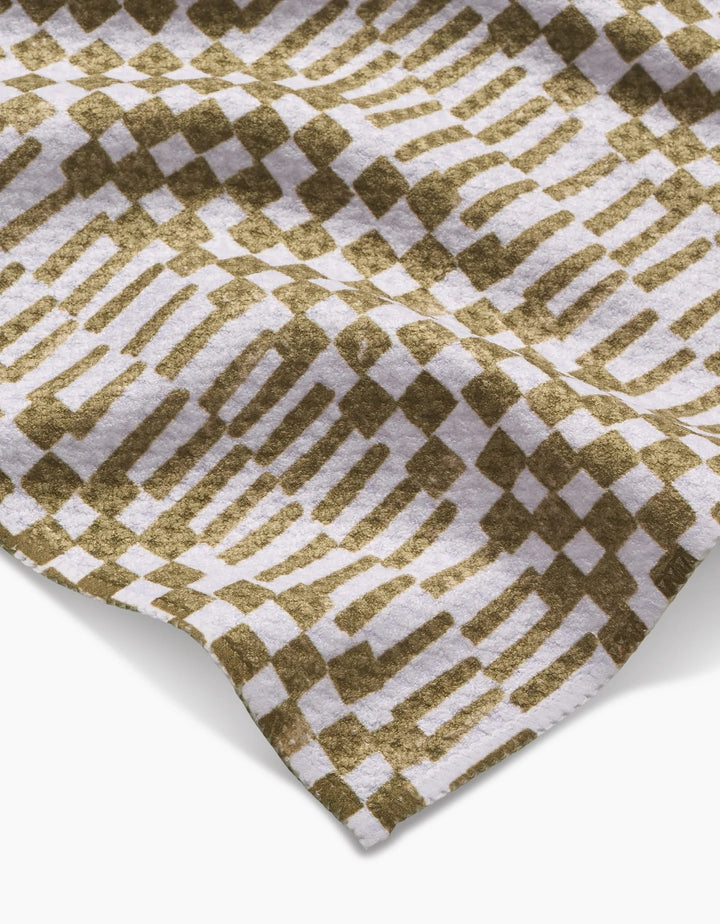 A close-up of Studio Jeannot’s Chutes Avocado bar towel showcases its Geoweave fabric in an olive green and white geometric pattern of alternating squares and elongated shapes, with a gently draped, textured appearance.