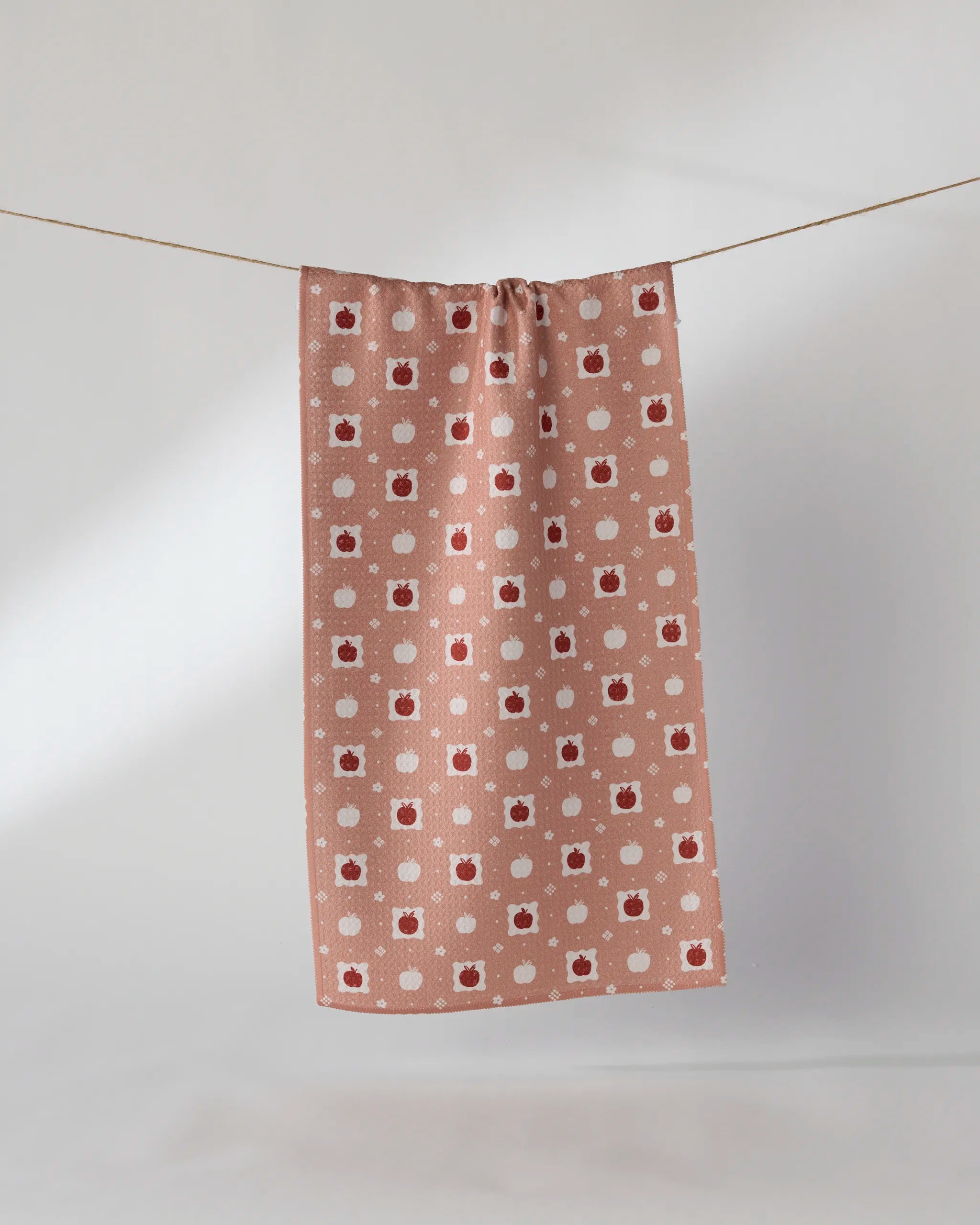 The Geometry "Cider Mill" towel, in pinkish-brown with a pattern of red apples and white dots, hangs on a string against a plain light background.