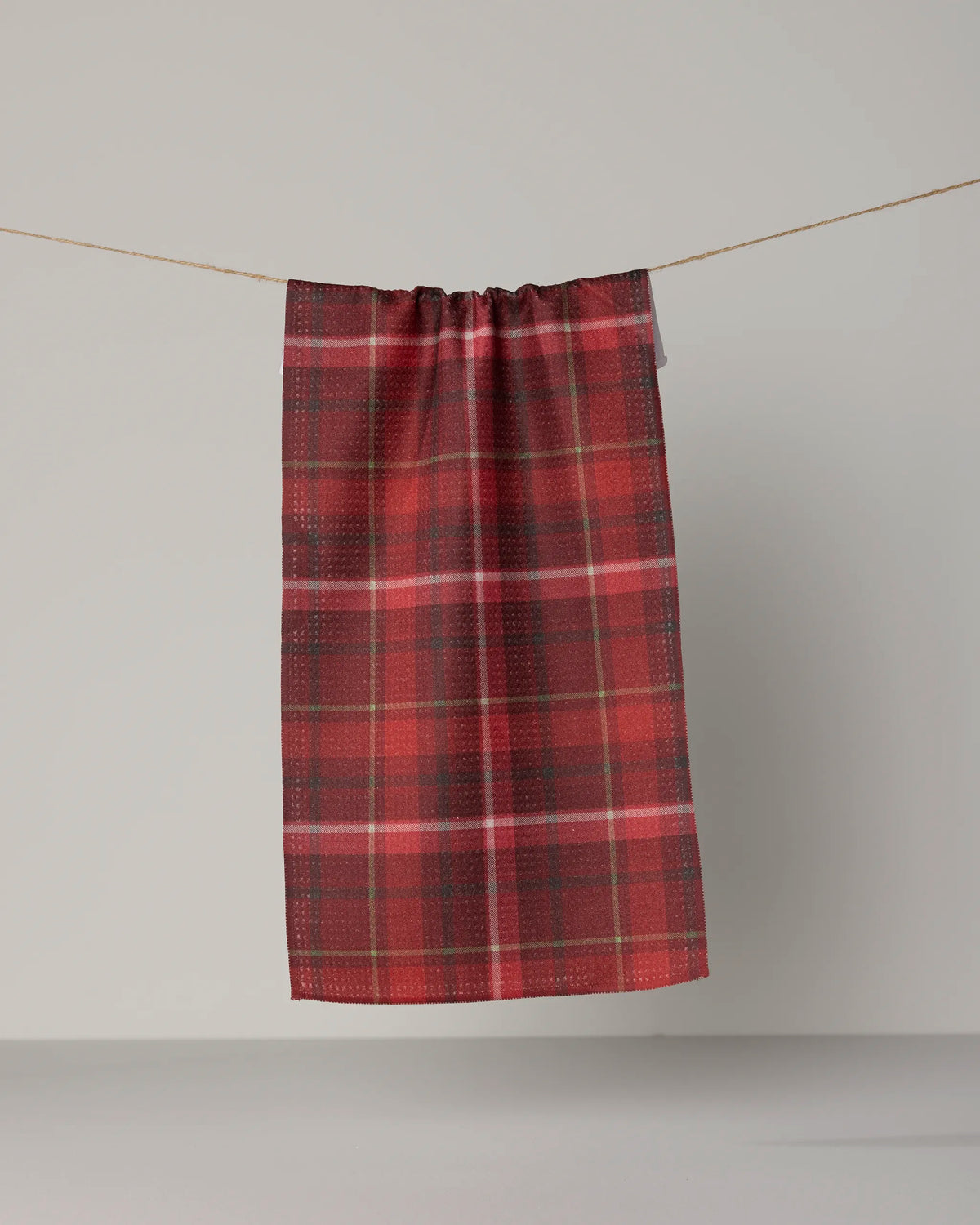 A Geometry Cinnamon Spice mini towel, featuring a red tartan pattern and crafted from Geoweave fabric, hangs from a string against a plain, light-colored background.