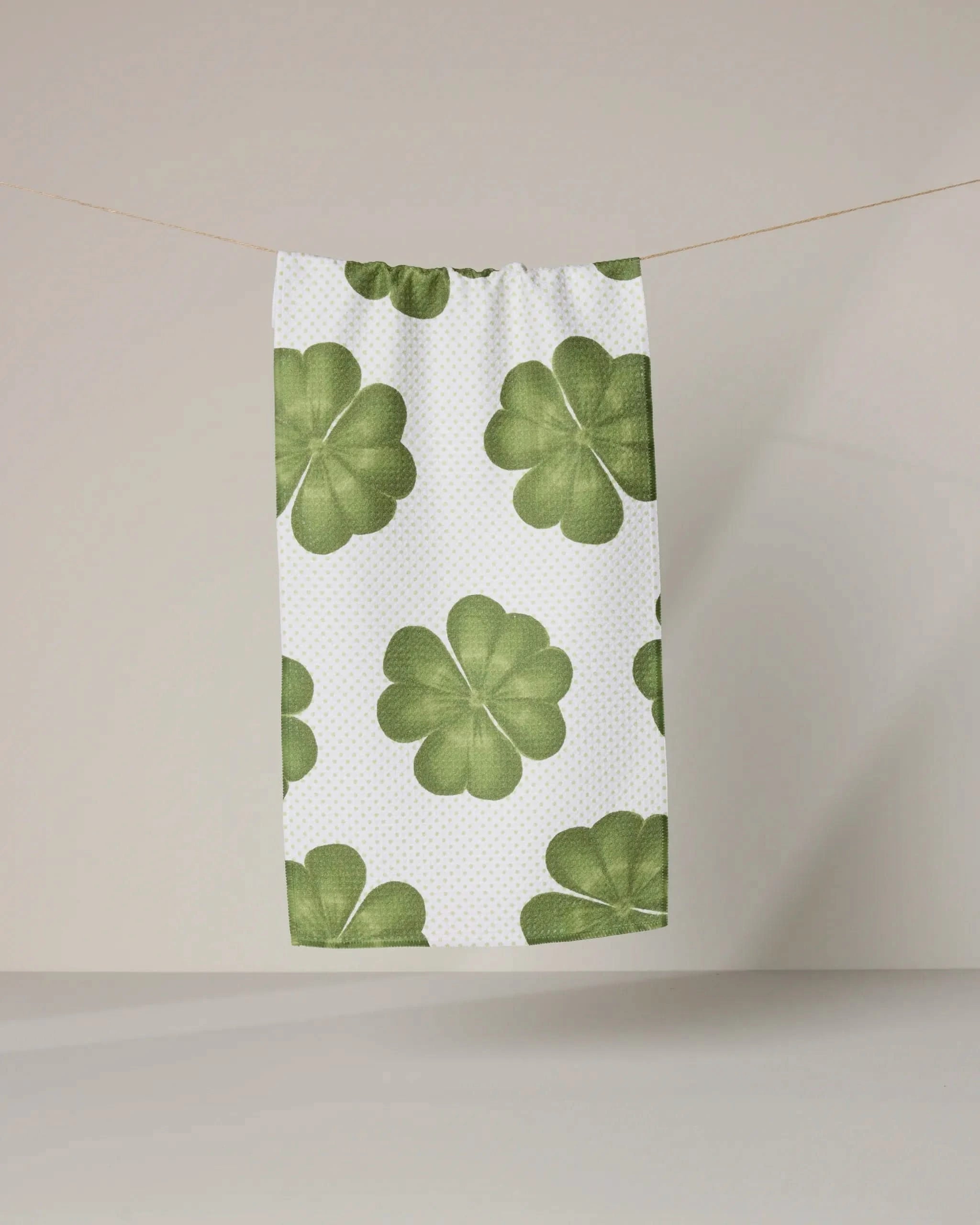 The Clover Maeve by Geometry, a white ultra-absorbent towel featuring large green four-leaf clovers, hangs on a clothesline against a plain light background.