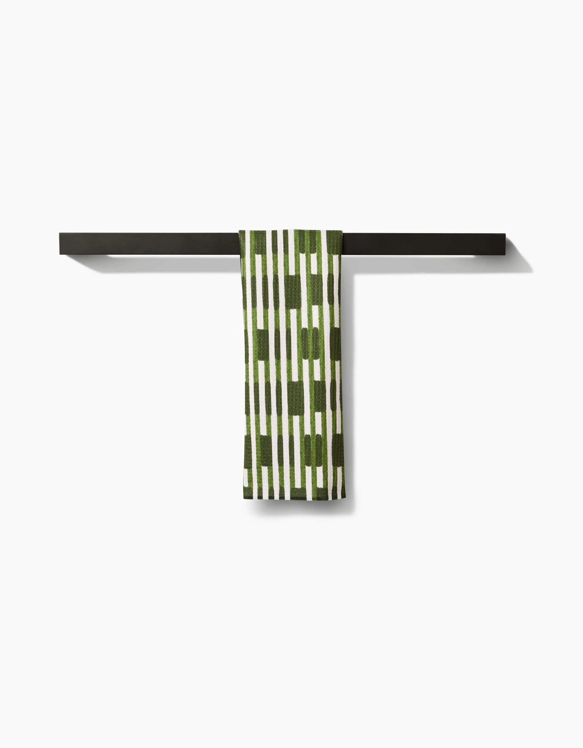 The GEOMETRY “Clover Stripe” quick-drying towel, featuring a green and white pattern, hangs neatly over a sleek black towel bar mounted on a white wall.