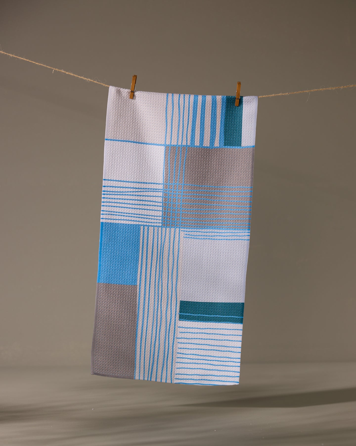 The Geometry Cool Blocks bar towel, featuring blue, gray, and white geometric patterns, hangs on a clothesline with two clothespins against a plain background.