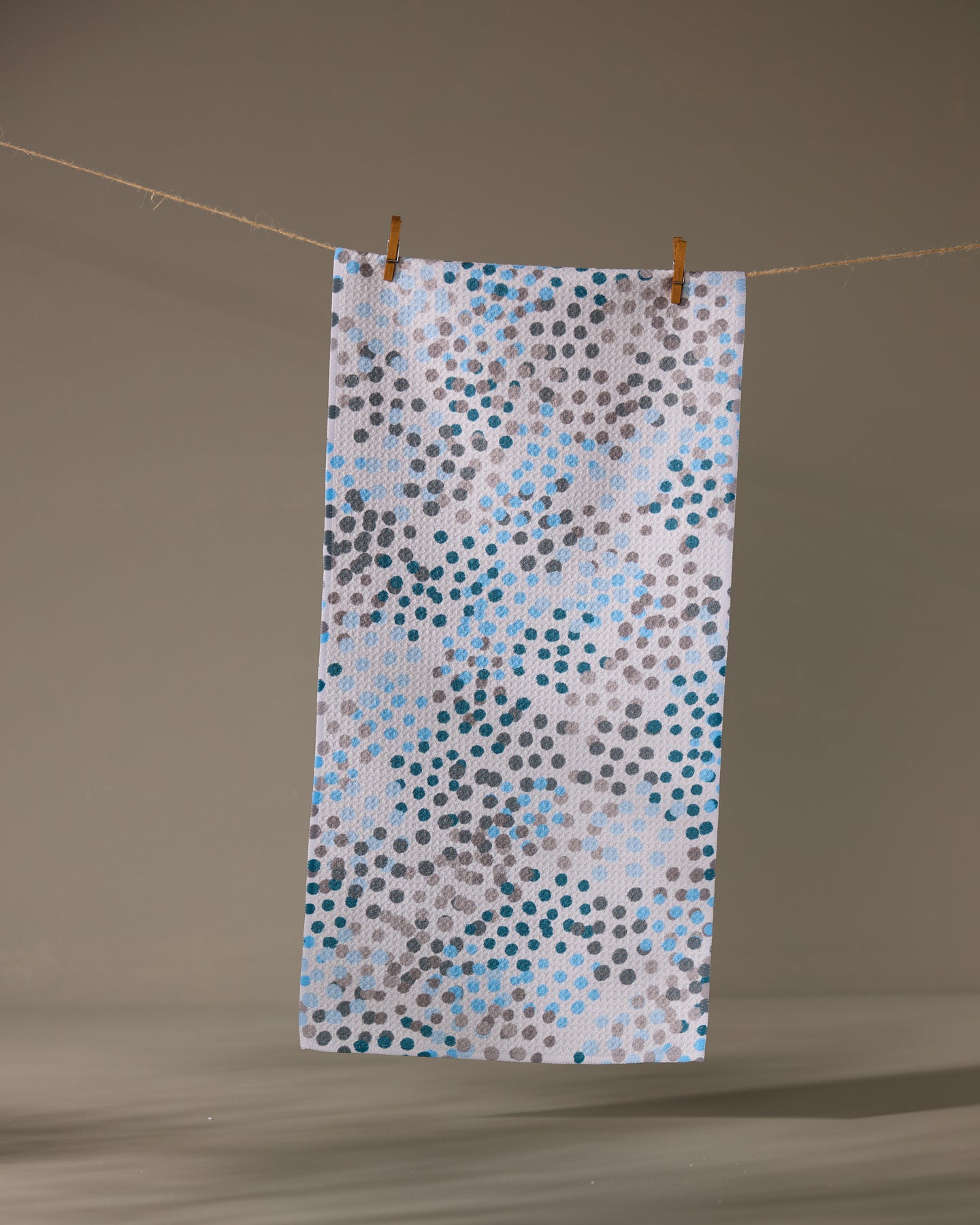 The Geometry Cool Dots recycled kitchen towel, white and rectangular with blue, grey, and black dot patterns, hangs from a string by two wooden clothespins against a simple background.
