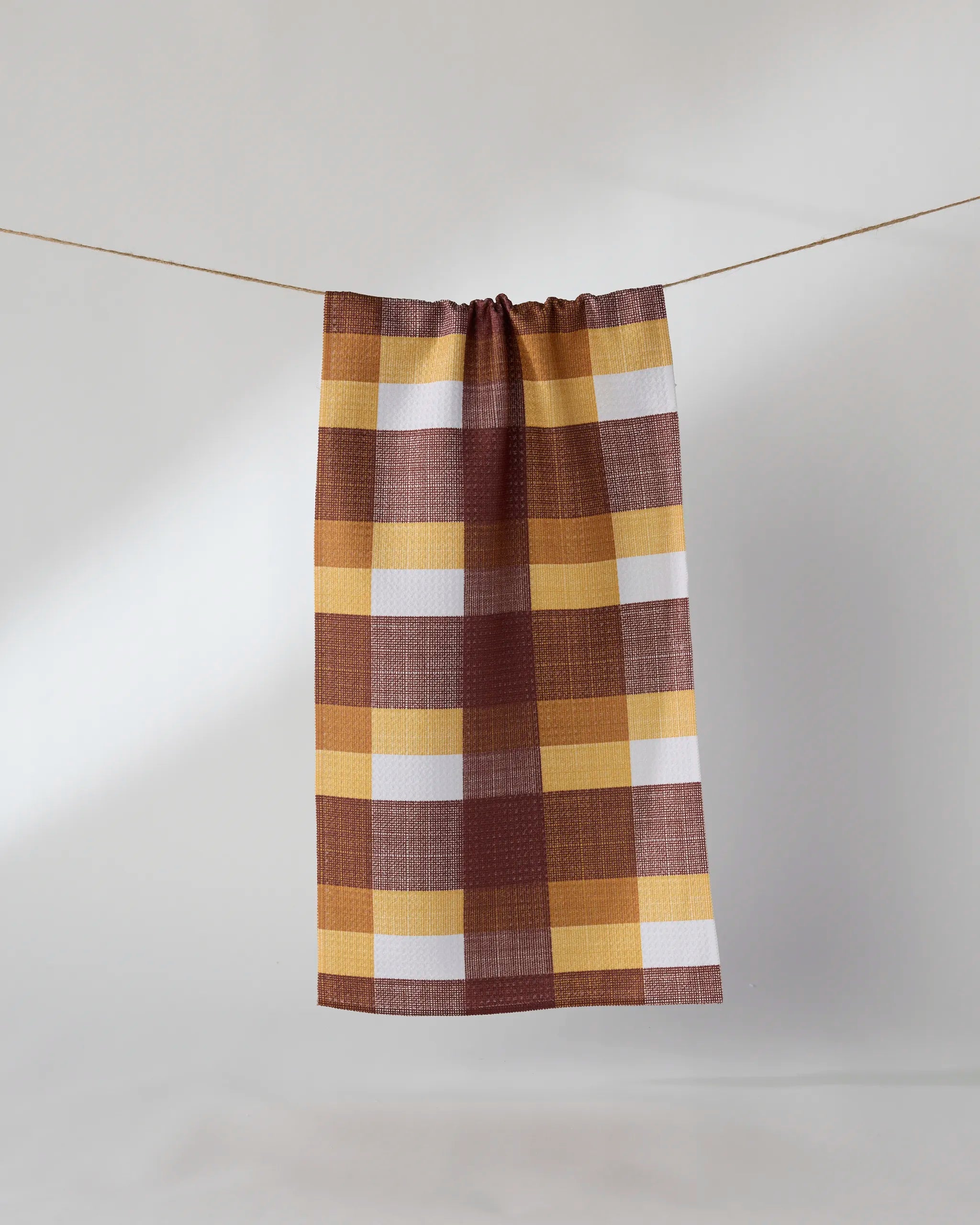 The Copper Grove bar towel by Geometry, featuring a brown, yellow, and white plaid design and made from recycled materials, hangs from a clothesline against a plain light background.
