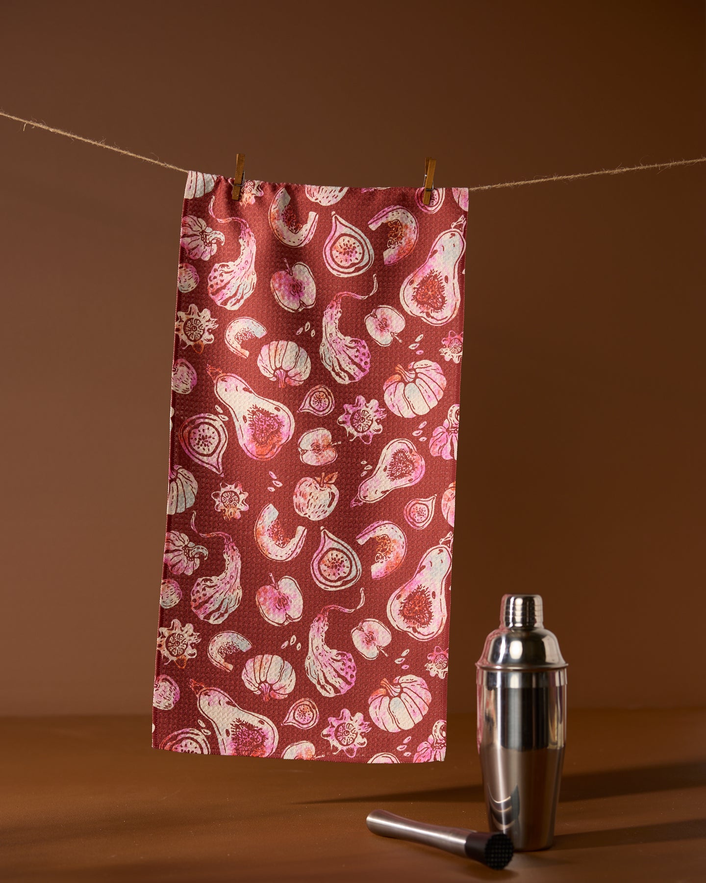 A red Geometry Cosecha ultra-absorbent bar towel with a white and pink vegetable pattern hangs on a string with clothespins. A metal cocktail shaker and muddler sit on a brown surface against a matching background.