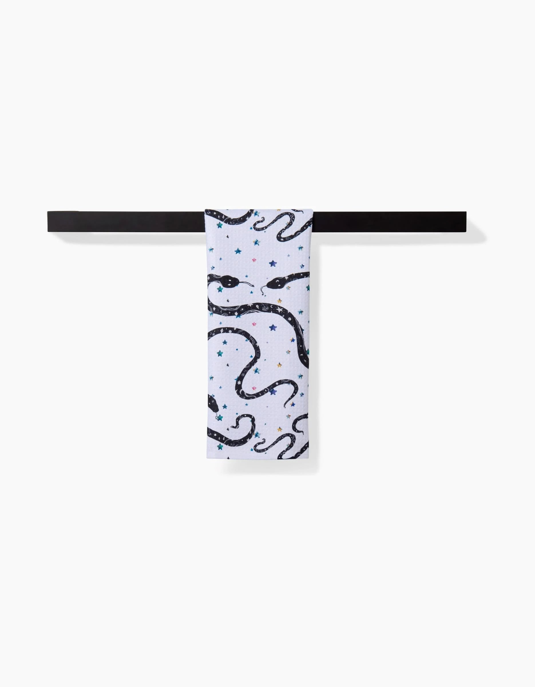 The “Cosmic Snakes” Bar Towel by Pascale Dilger, crafted from Geoweave™ fabric, features black snake and blue star designs, draped over a sleek horizontal black bar against a plain white background.