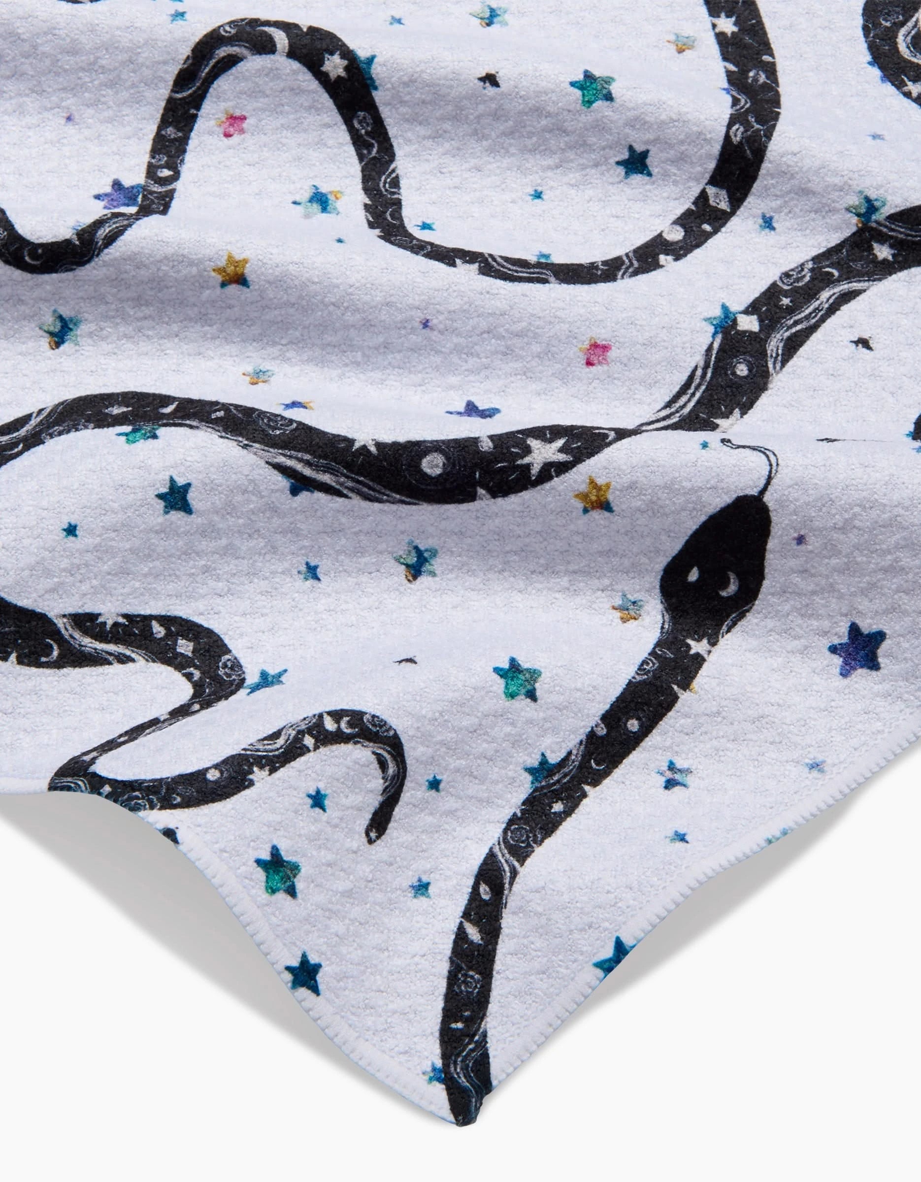A close-up of Pascale Dilgers Cosmic Snakes Bar Towel, crafted from Geoweave™ fabric, shows a black snake with intricate patterns and vibrant blue and purple stars. The snake’s head appears near a folded corner of the towel.