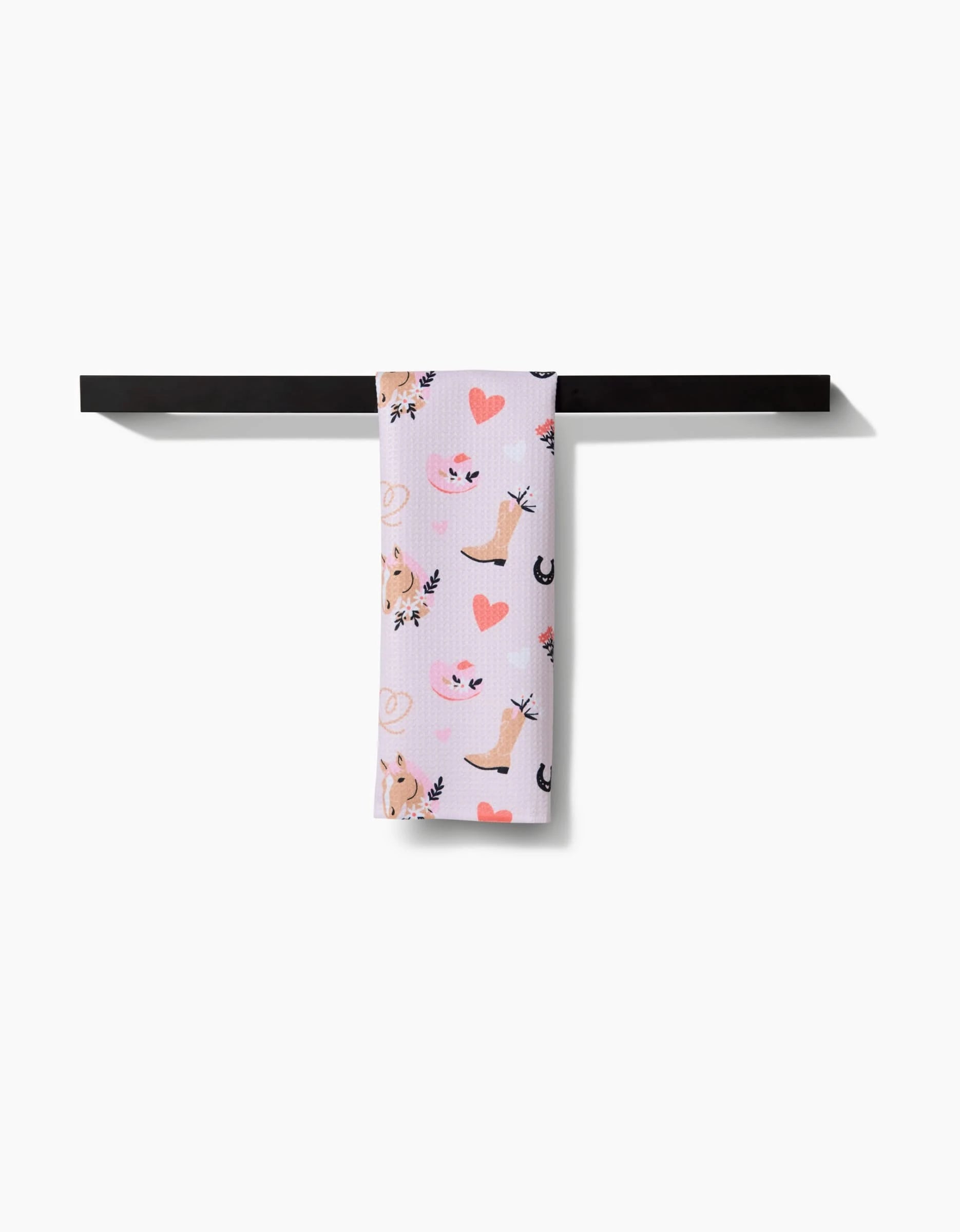 The Cowgirl Love towel by Amberleigh Adoff, featuring a pink cat, heart, and leaf print on ultra-absorbent white fabric, is neatly draped over a sleek black towel bar against a white background.