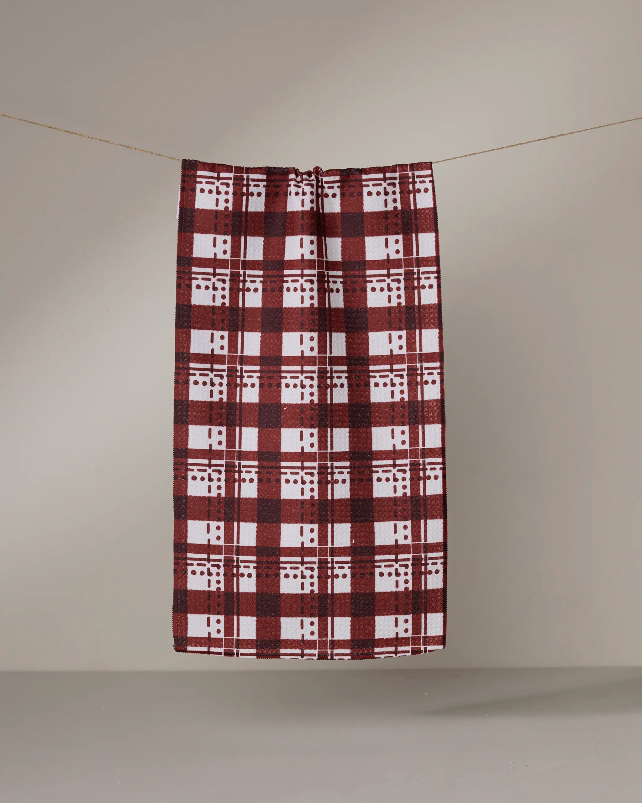 The Geometry Cranberry Plaid bar towel hangs on a thin clothesline against a plain, light grey background.