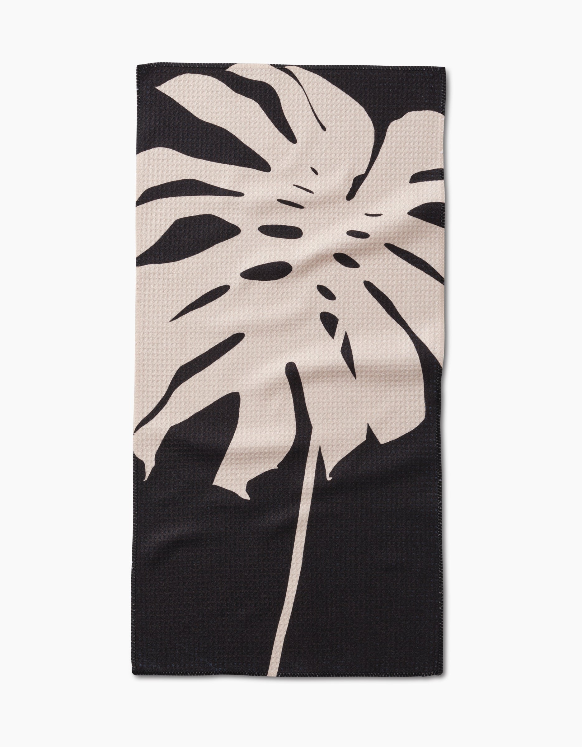The Cream Monstera Bar Towel by PrintedPlant features a large beige monstera leaf on dark Geoweave fabric, offering superior absorbency and style.