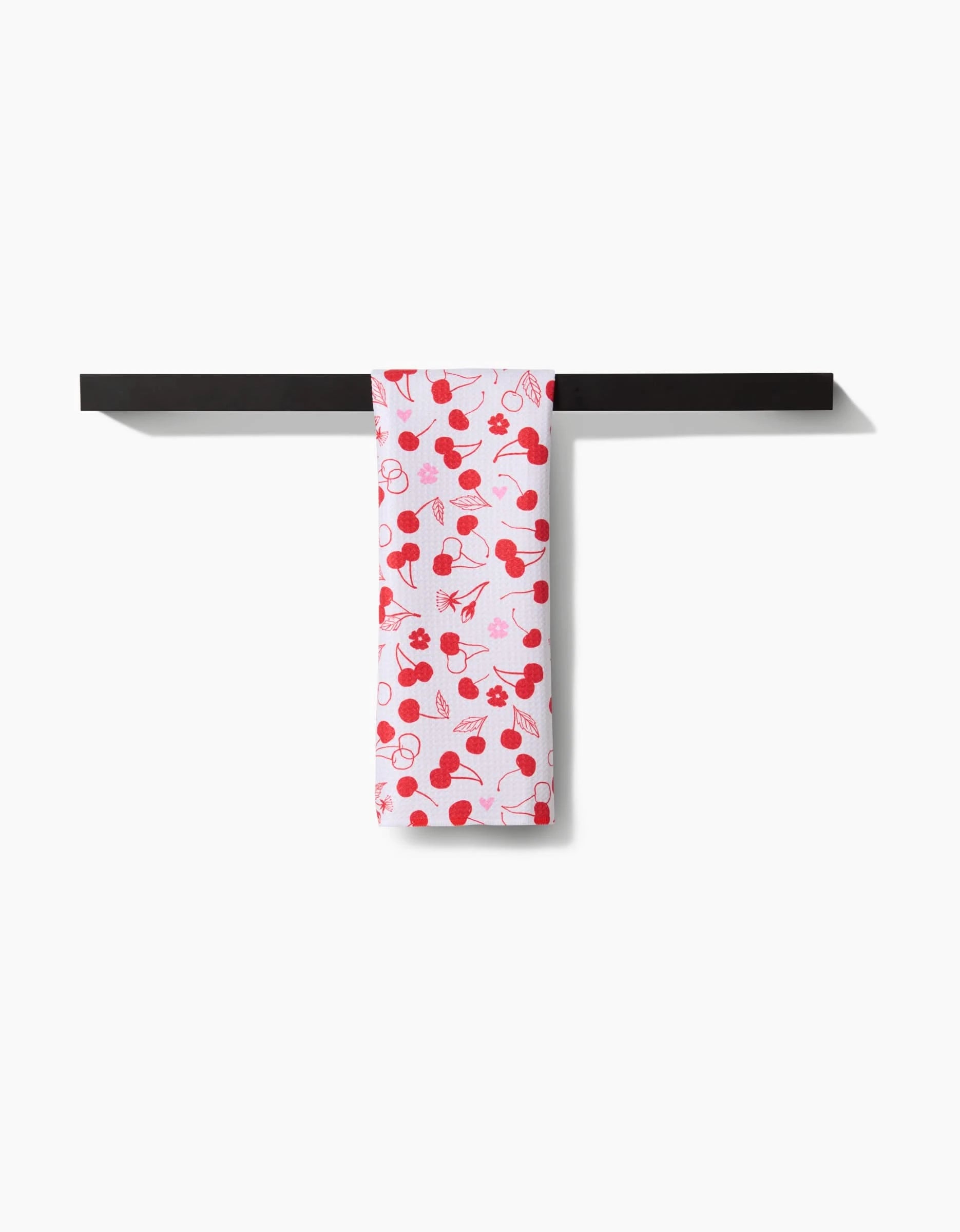 The Cute Cherry bar towel by Elana Gabrielle, featuring a red cherry pattern on white and made from ultra-absorbent recycled materials, is displayed draped over a minimalist black bar against a plain white background.