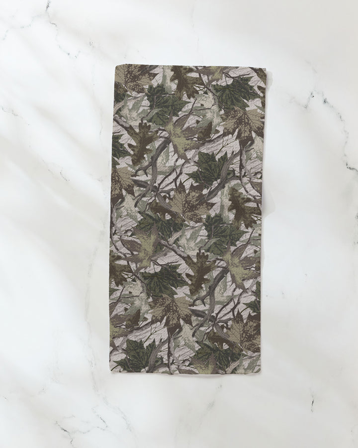 The Dad Camo by Geometry is a rectangular bar towel featuring a green and brown leaf camouflage pattern, made from recycled materials, shown on a white marble surface.