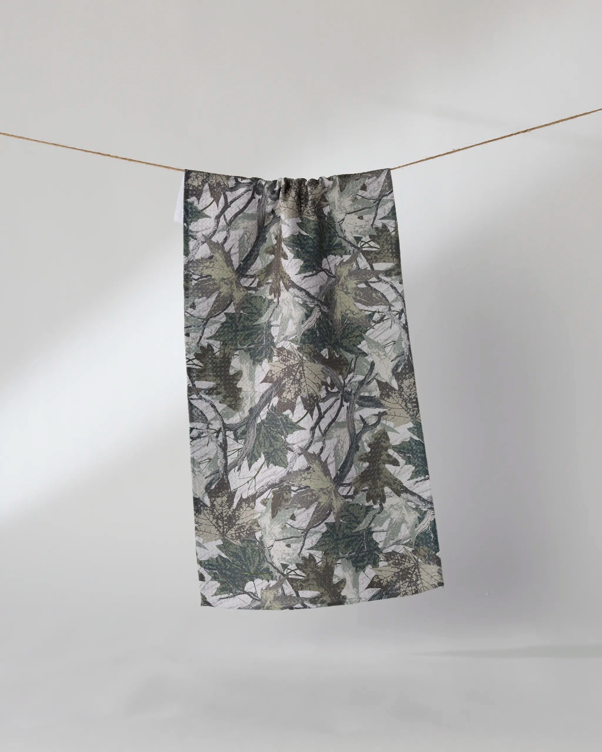 The Geometry "Dad Camo" towel, featuring a green and gray leafy camouflage pattern and ultra-absorbent rectangular design, hangs from a clothesline against a plain light background.