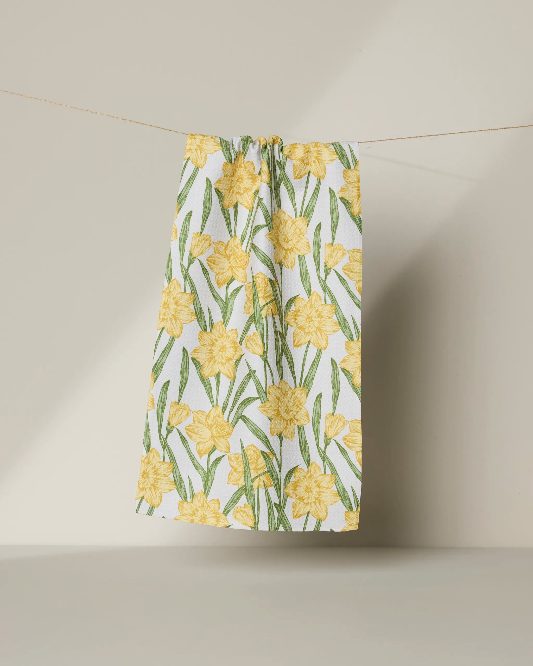 The Geometry Daffodil Darling towel, featuring ultra-absorbent fabric and a yellow daffodil and green leaf pattern, hangs on a string against a soft, light backdrop.