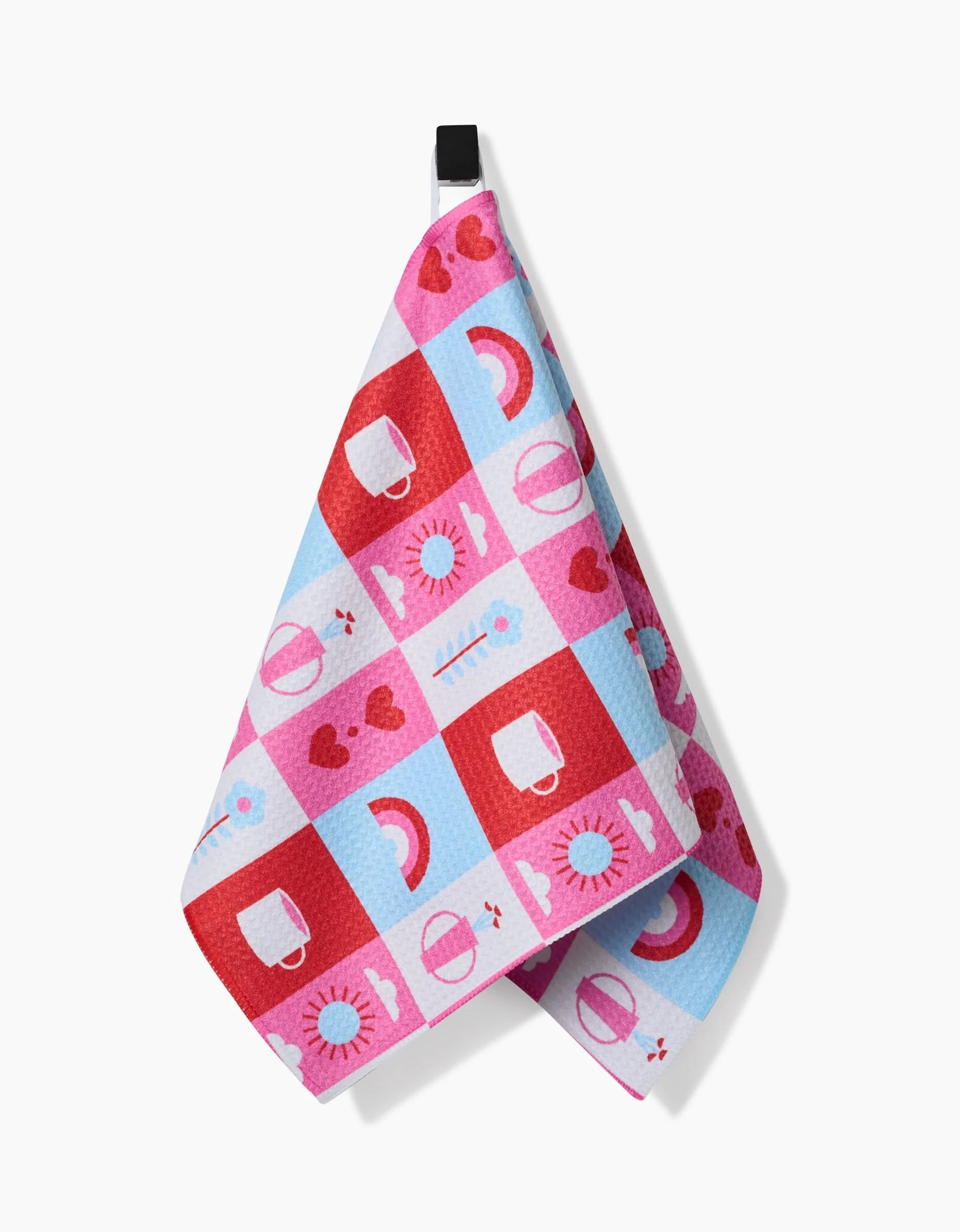 The Daily Icons Bar Towel by Laura Supnik is made from recycled materials and features a red, pink, and blue checkered pattern with cups, hearts, rainbows, and sun motifs. It hangs from a black loop against a white background.