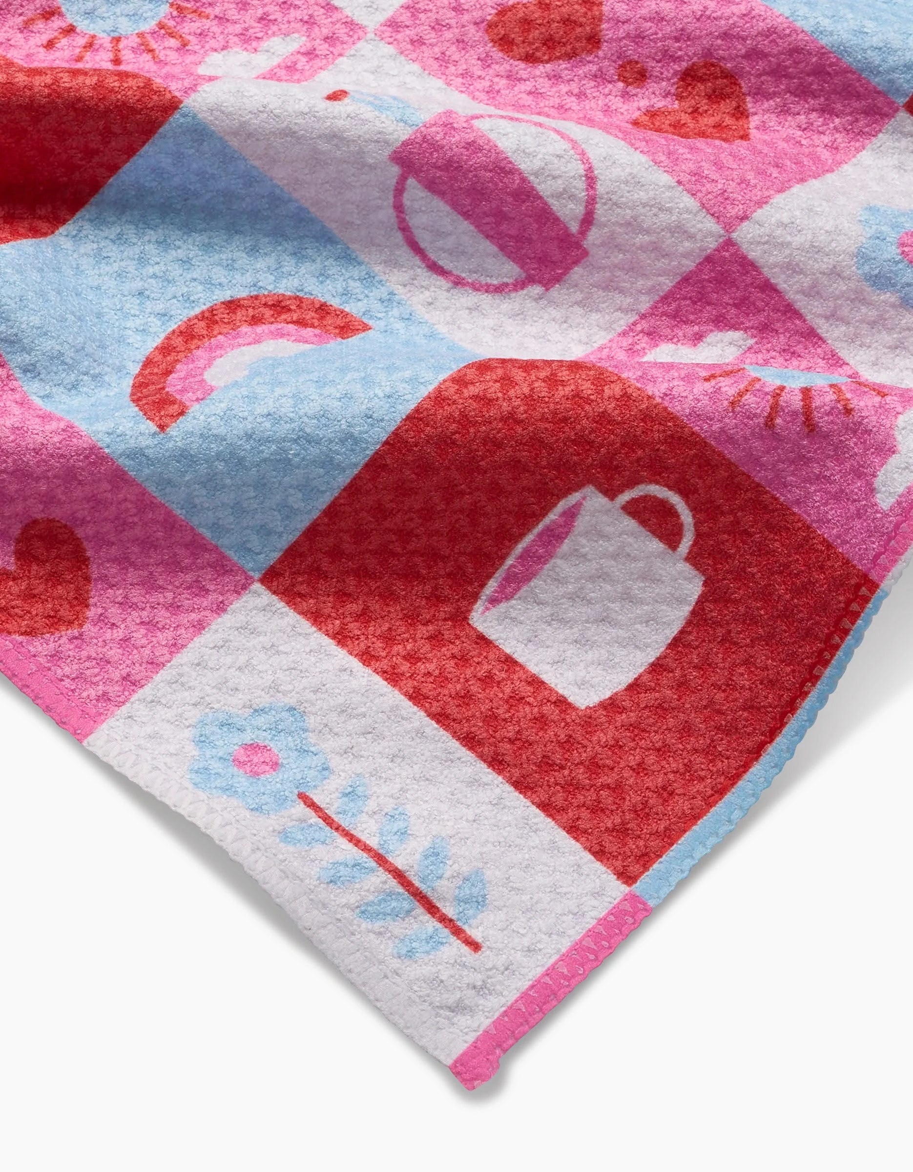A close-up of Laura Supnik’s Daily Icons bar towel in pink, red, and blue checks with playful mugs, flowers, rainbows, hearts, and suns—crafted from recycled materials for a sustainable touch.