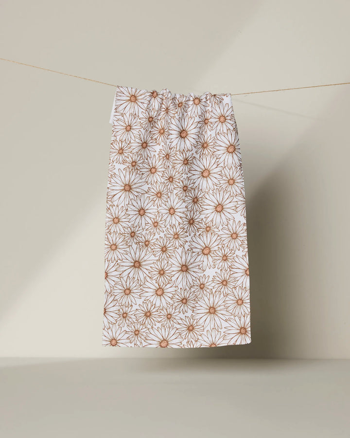 The Daisy Trail kitchen towel by Geometry, made from recycled materials, features a brown daisy flower pattern on white and hangs from a thin string against a neutral beige background.