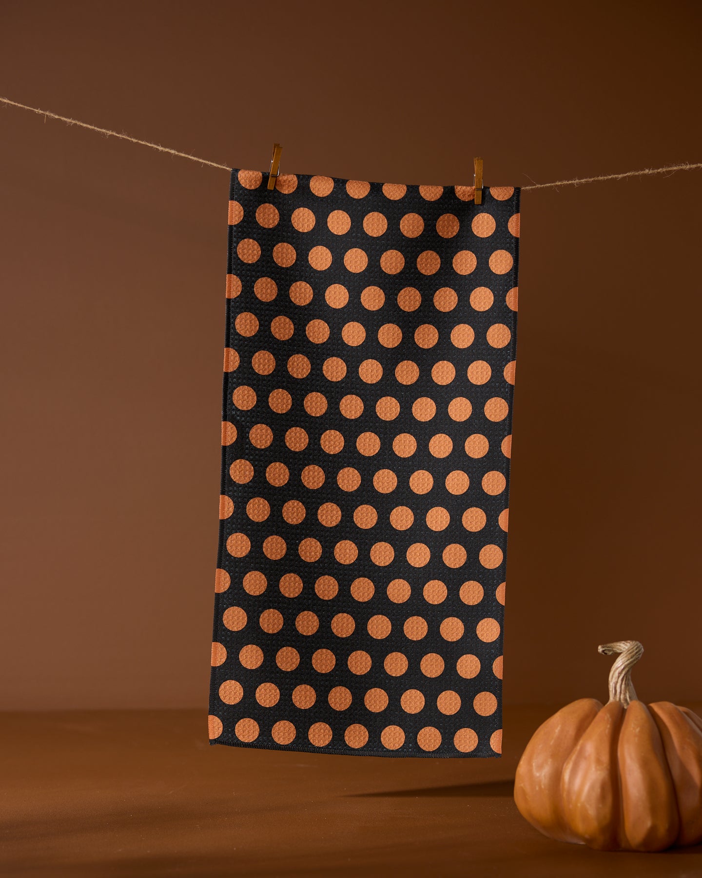 The Geometry Dot Noir towel, in black with orange polka dots, hangs on a clothesline with two clothespins above an orange pumpkin decoration on a brown surface, set against a brown background.