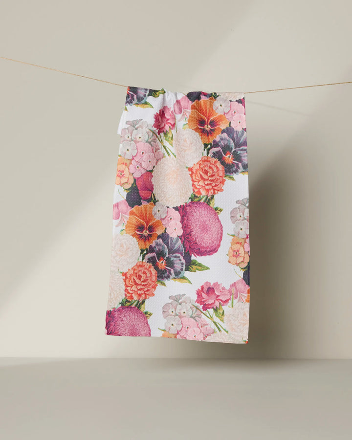 The Geometry Ecstatic Blooms towel, featuring vibrant pink, orange, purple, and white floral patterns, hangs from a string against a plain light background.