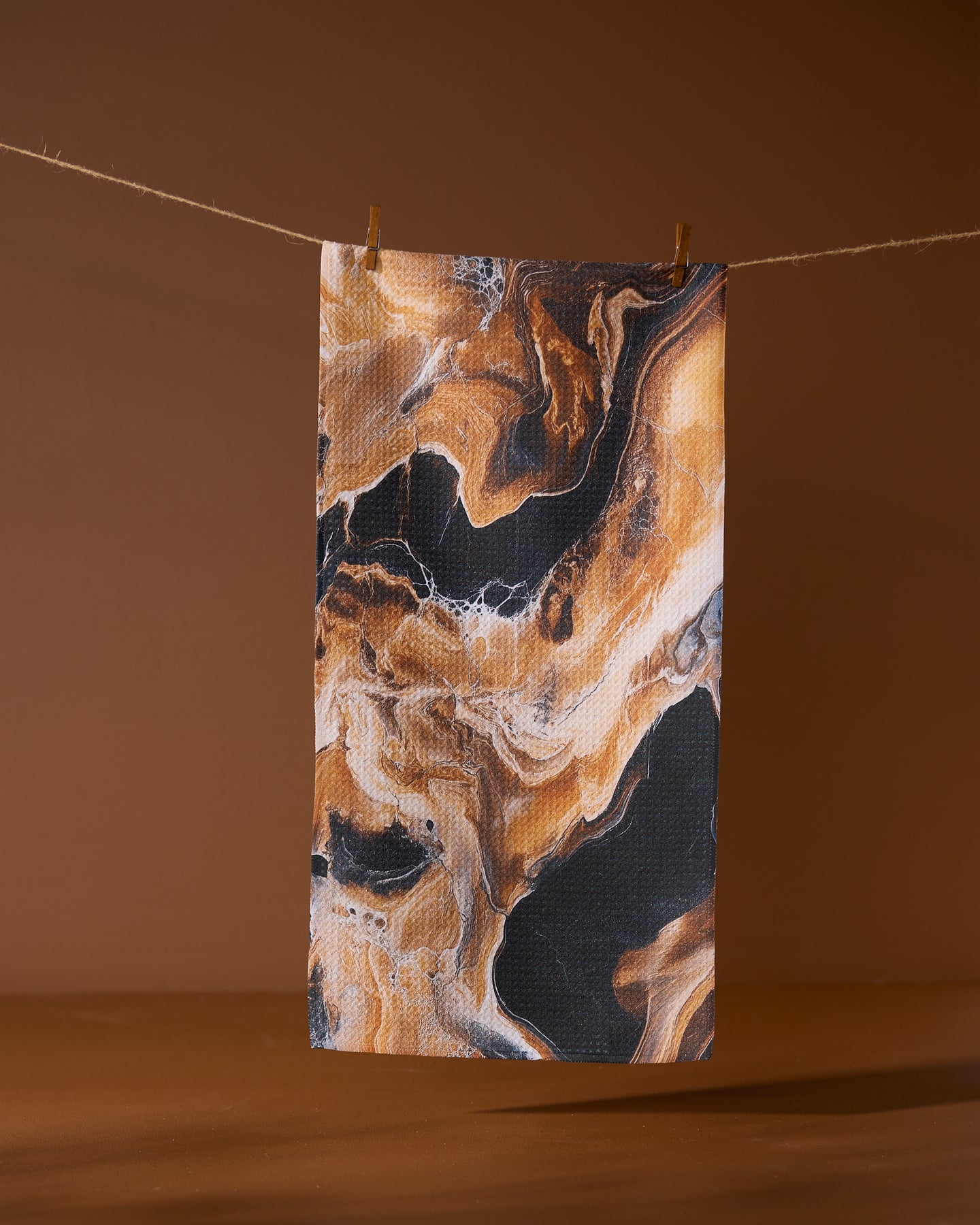 The Geometry Emperador rectangular recycled kitchen towel, featuring a black, brown, and beige marbled pattern, hangs from a string with two clothespins against a solid brown background.