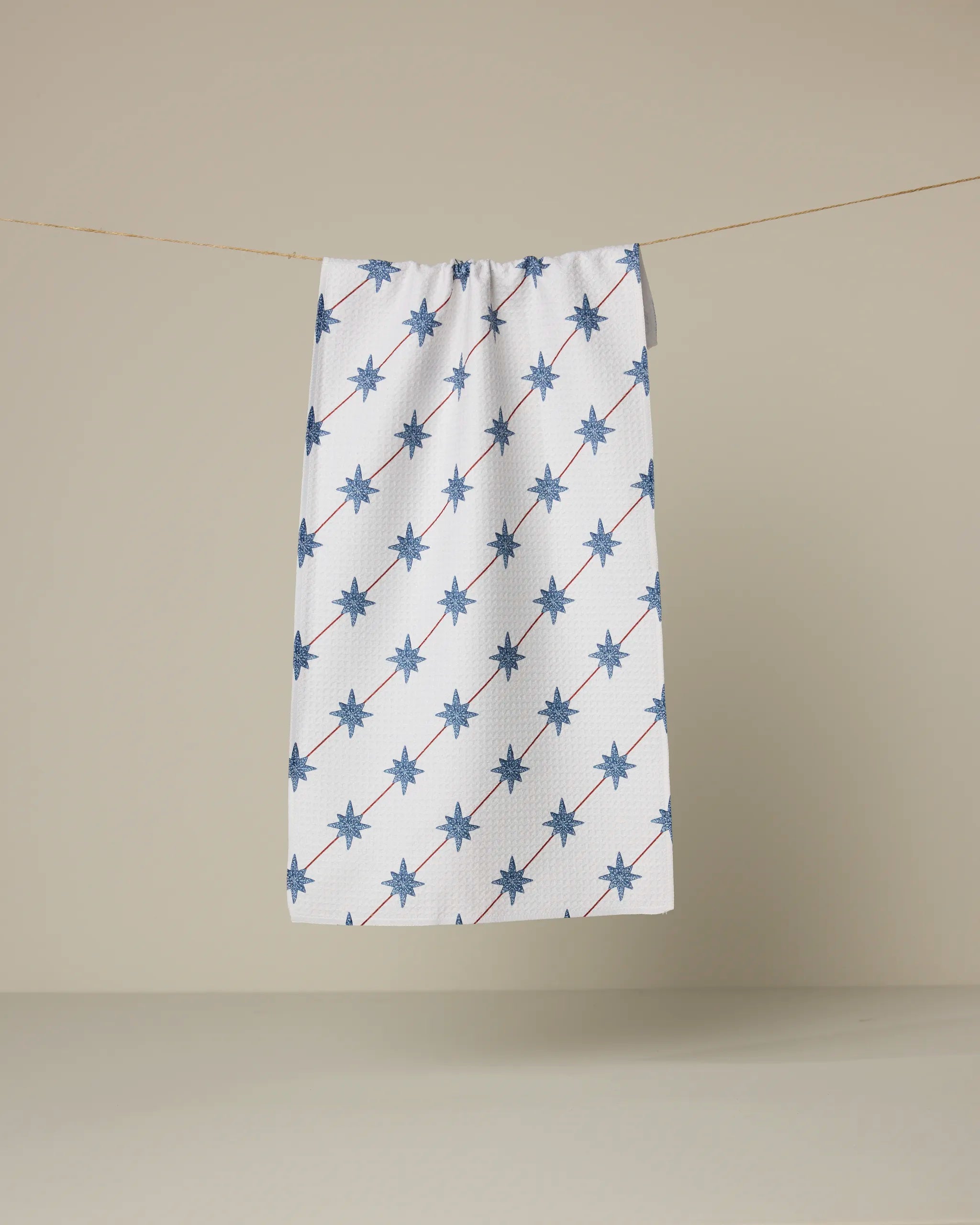 The Estrella Norte towel by Geometry, featuring blue eight-pointed stars in diagonal lines on a white background, hangs on a clothesline against plain beige.
