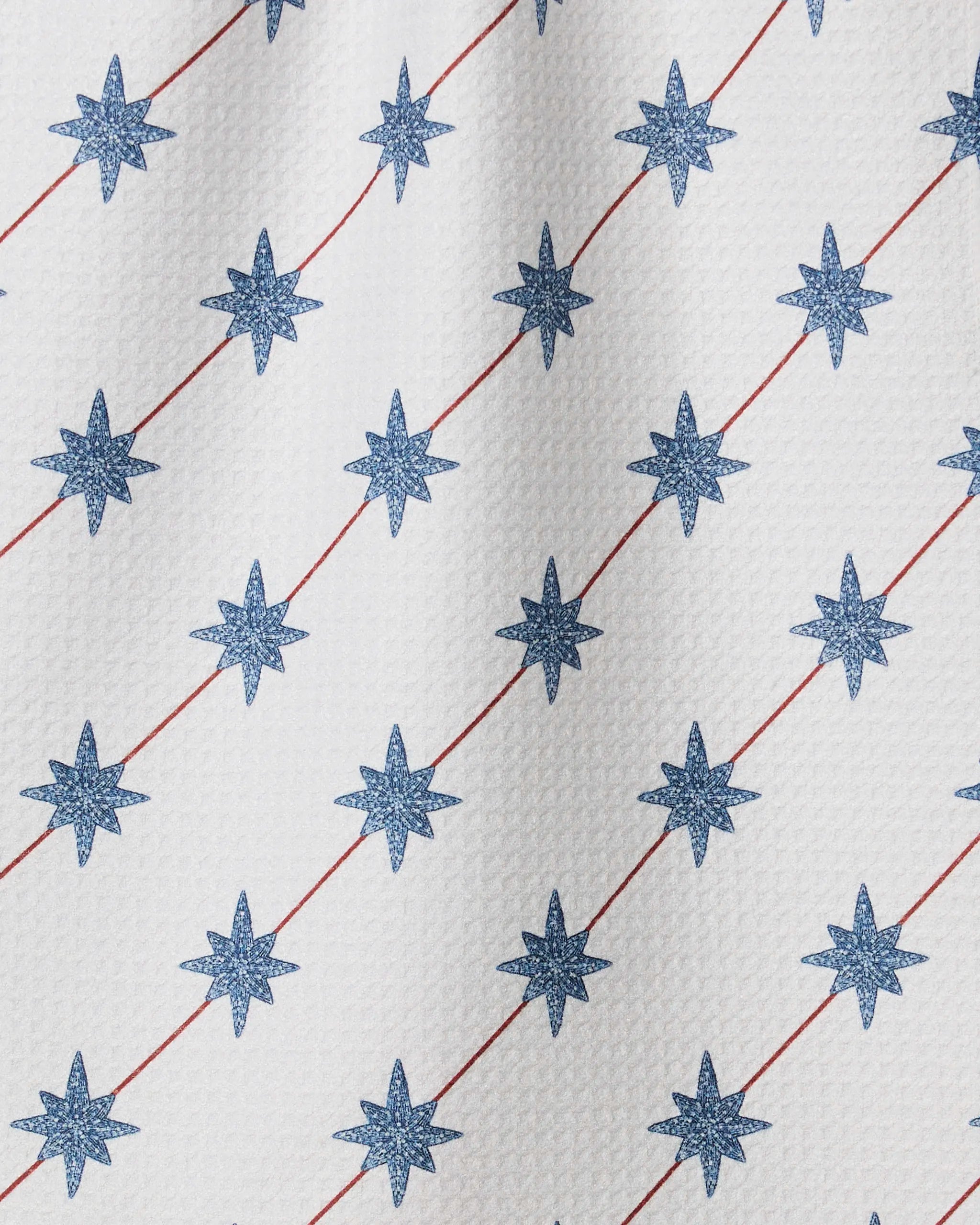 The Estrella Norte towel by Geometry is white and quick-drying, featuring a textured surface with blue eight-pointed stars in diagonal rows, intersected by thin red diagonal lines.