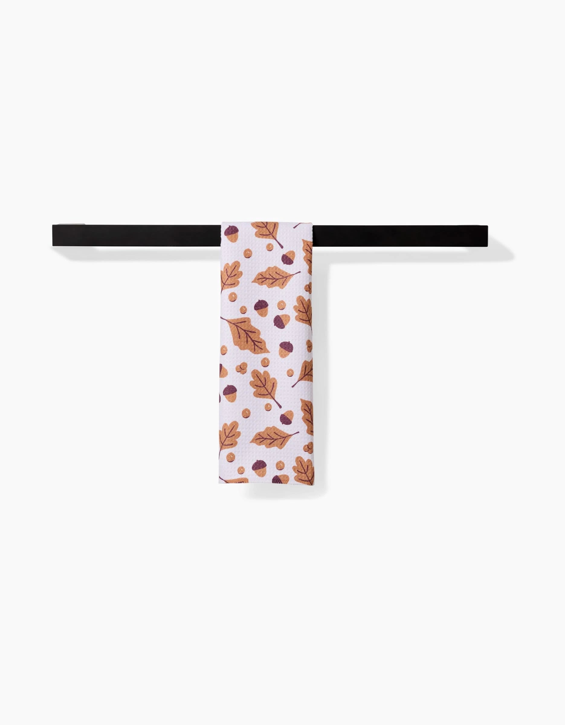 The Falling For Fall kitchen towel by Charlie Rowan Designs, featuring brown leaves and acorns, hangs neatly on a black towel bar against a plain white background.