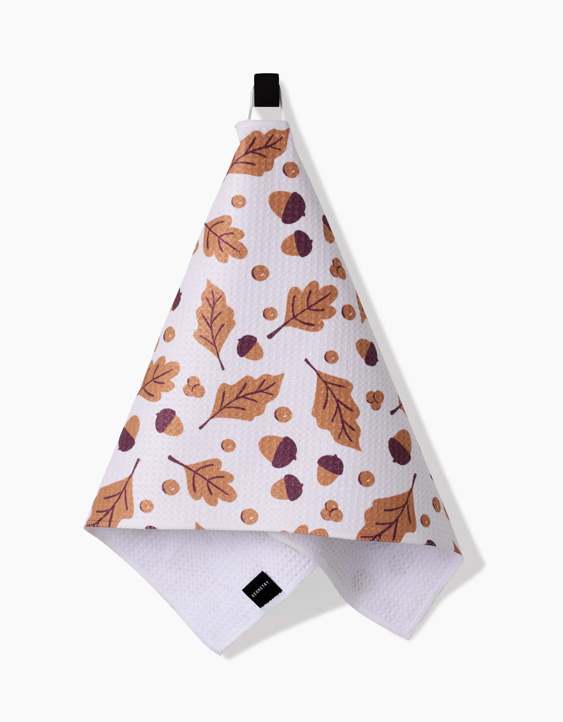 The Falling For Fall kitchen bar towel by Charlie Rowan Designs is a quick-drying, white textured towel with an autumn pattern of brown and orange leaves, acorns, and berries, and hangs from a black loop.