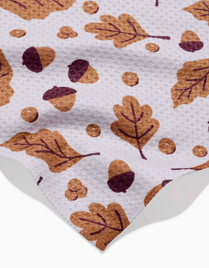 Falling For Fall by Charlie Rowan Designs is a white textured recycled kitchen towel featuring brown and dark purple acorns, oak leaves, and berries, with part of the cloth edge visible.