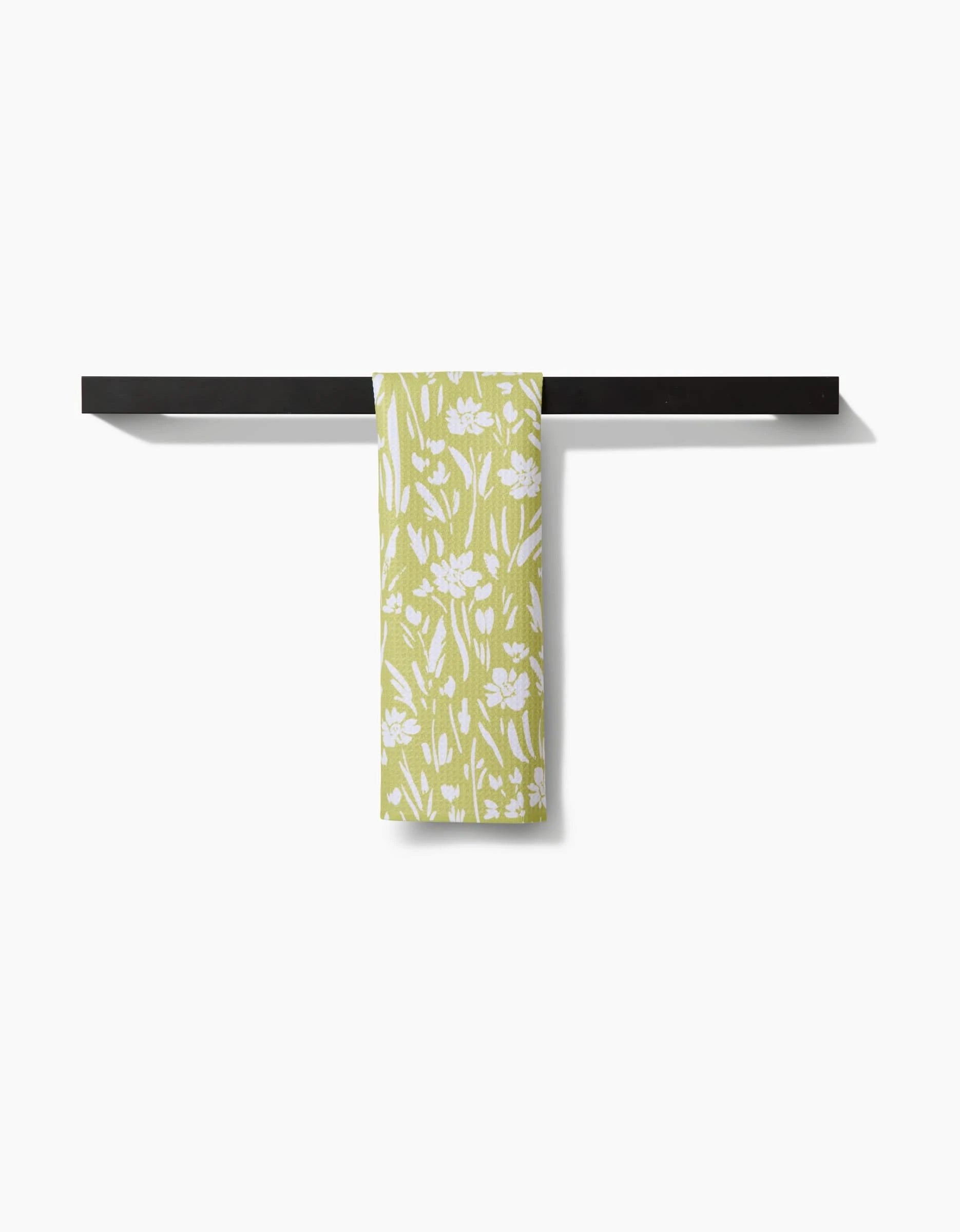 The Field of Wildflowers bar towel by Ashley Celena features a green hue with a white floral pattern, made from recycled materials, and is shown folded over a black bar on a plain white background.