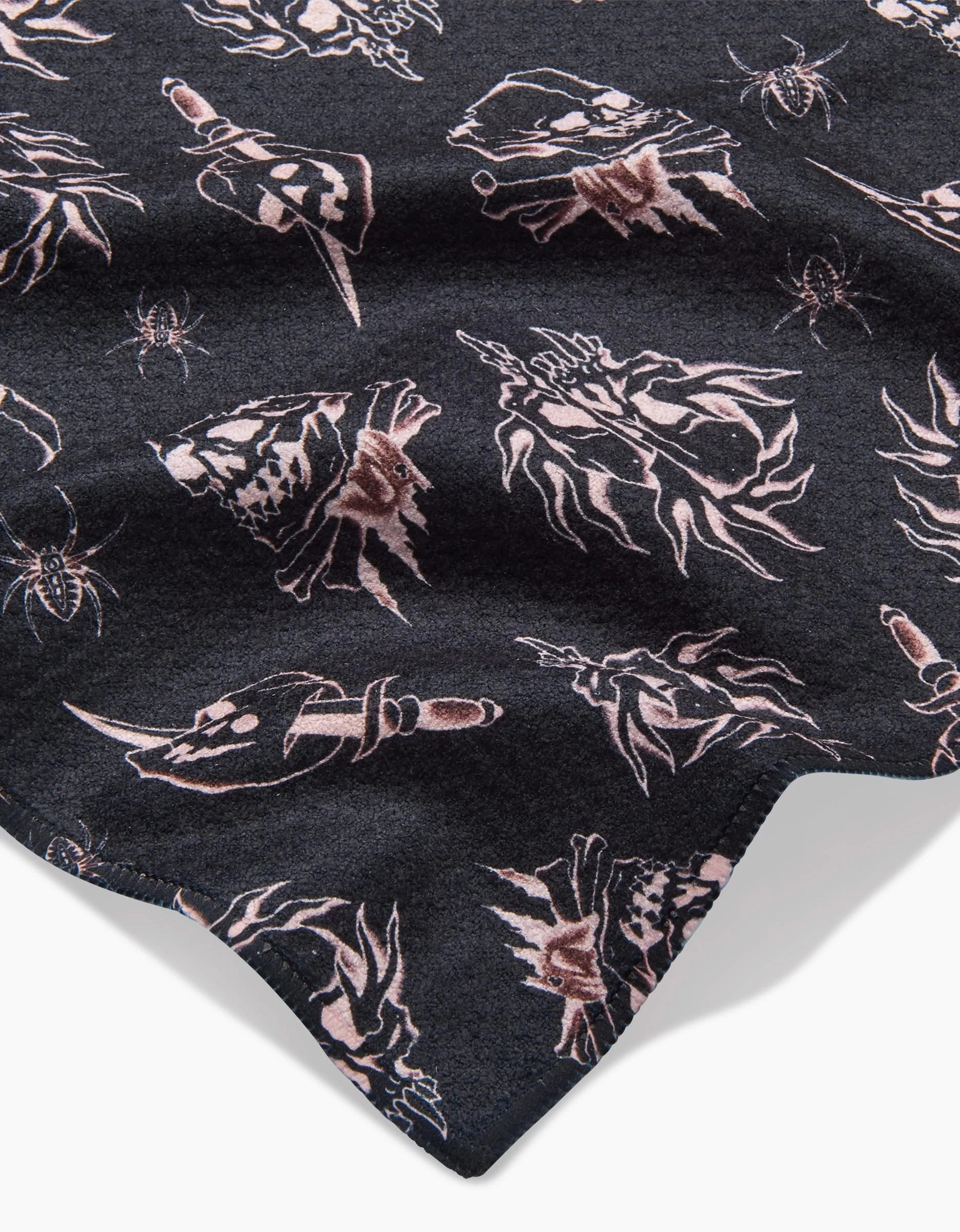 A close-up of Cole Strem’s Fiery Skulls ultra-absorbent black bar towel, made from recycled materials, with pinkish skulls, daggers, and flame motifs. The textured fabric surface reveals subtle folds.