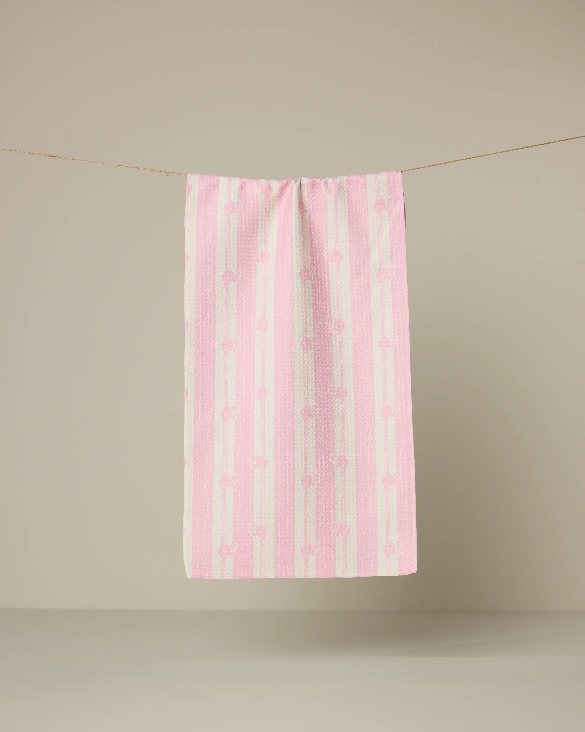 The Geometry Flamingo Stripe Pink towel, featuring pink and white stripes with a bow pattern, hangs from a clothesline against a plain beige background.