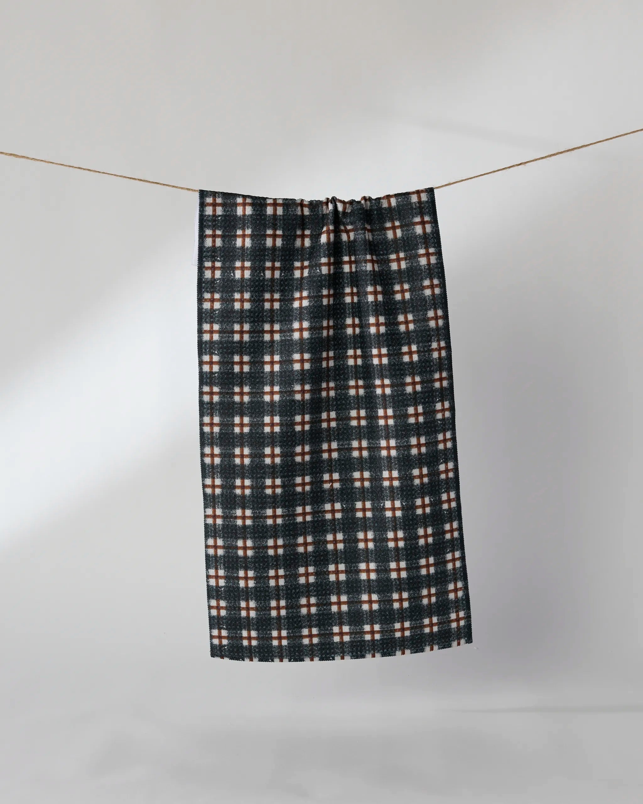 The Geometry Flint bar towel, featuring dark plaid with white, red, and blue stripes, hangs on a string against a plain light gray background.