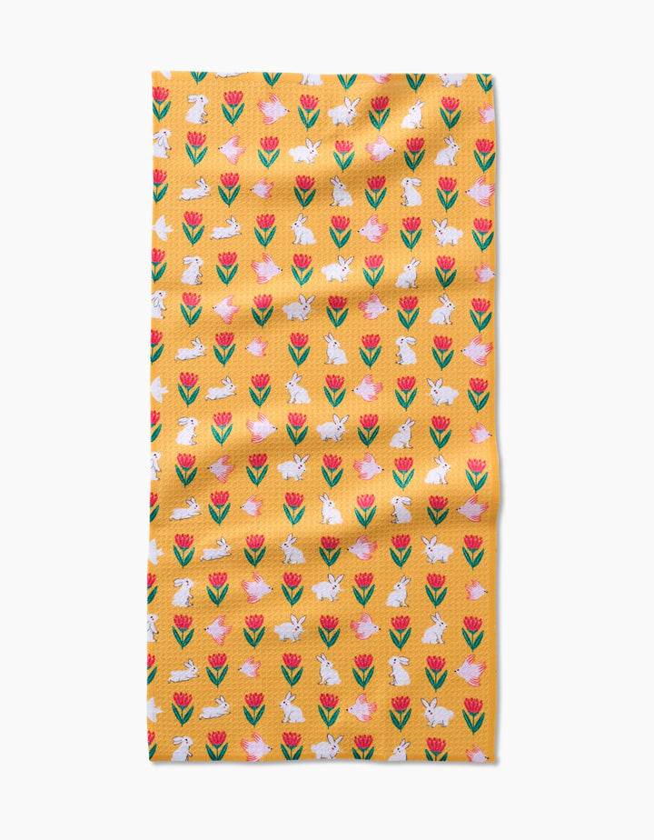 The Floral Bunny Parade kitchen towel by Ceyda Alasar features a yellow recycled fabric with red and pink tulips and white bunnies in a repeating pattern, shown slightly wrinkled on a white background.