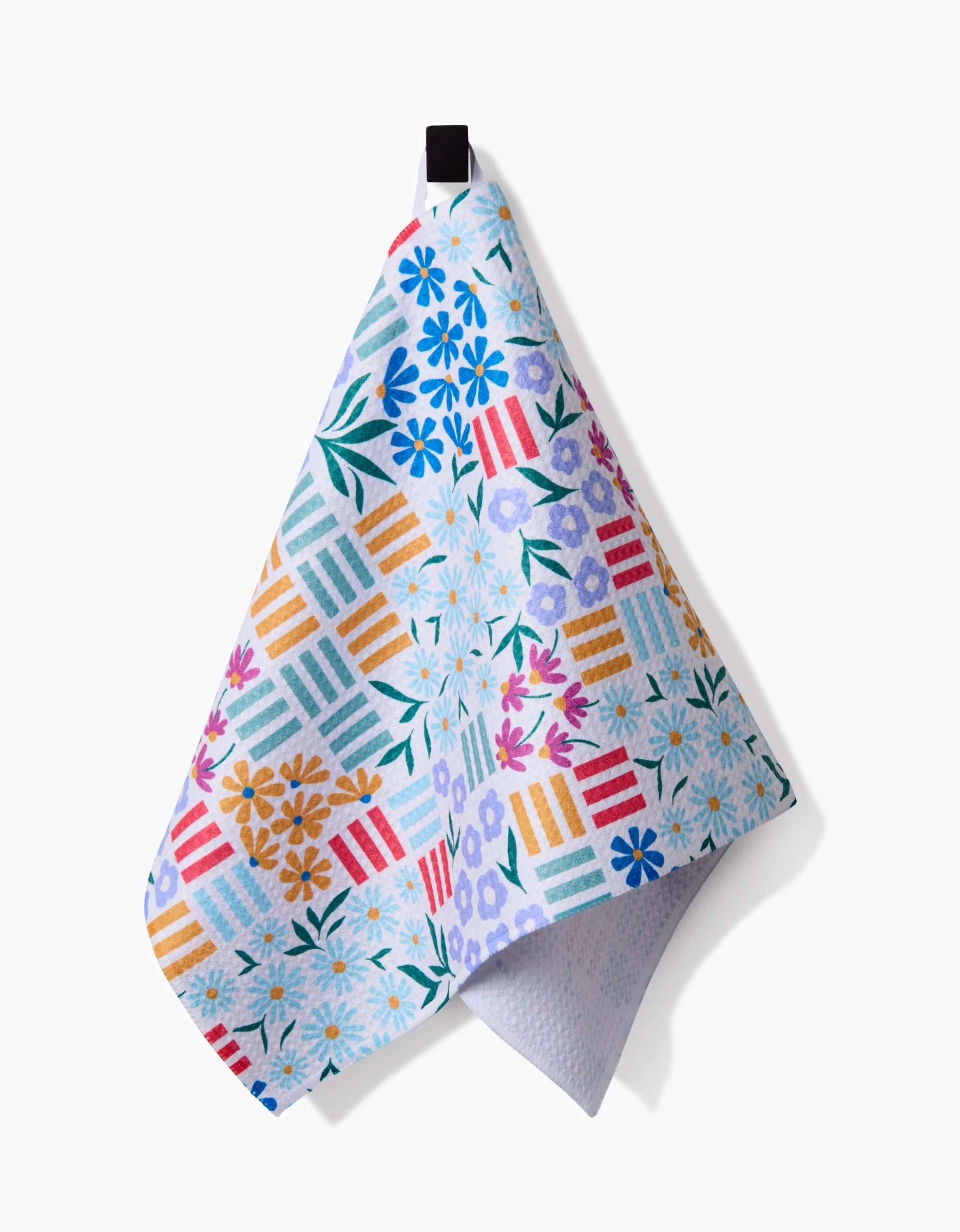 The Floral Garden dish towel by Charlie Rowan Designs hangs on a hook, made from Geoweave fabric and featuring blue, purple, yellow, and orange flowers with geometric stripes on a white background.