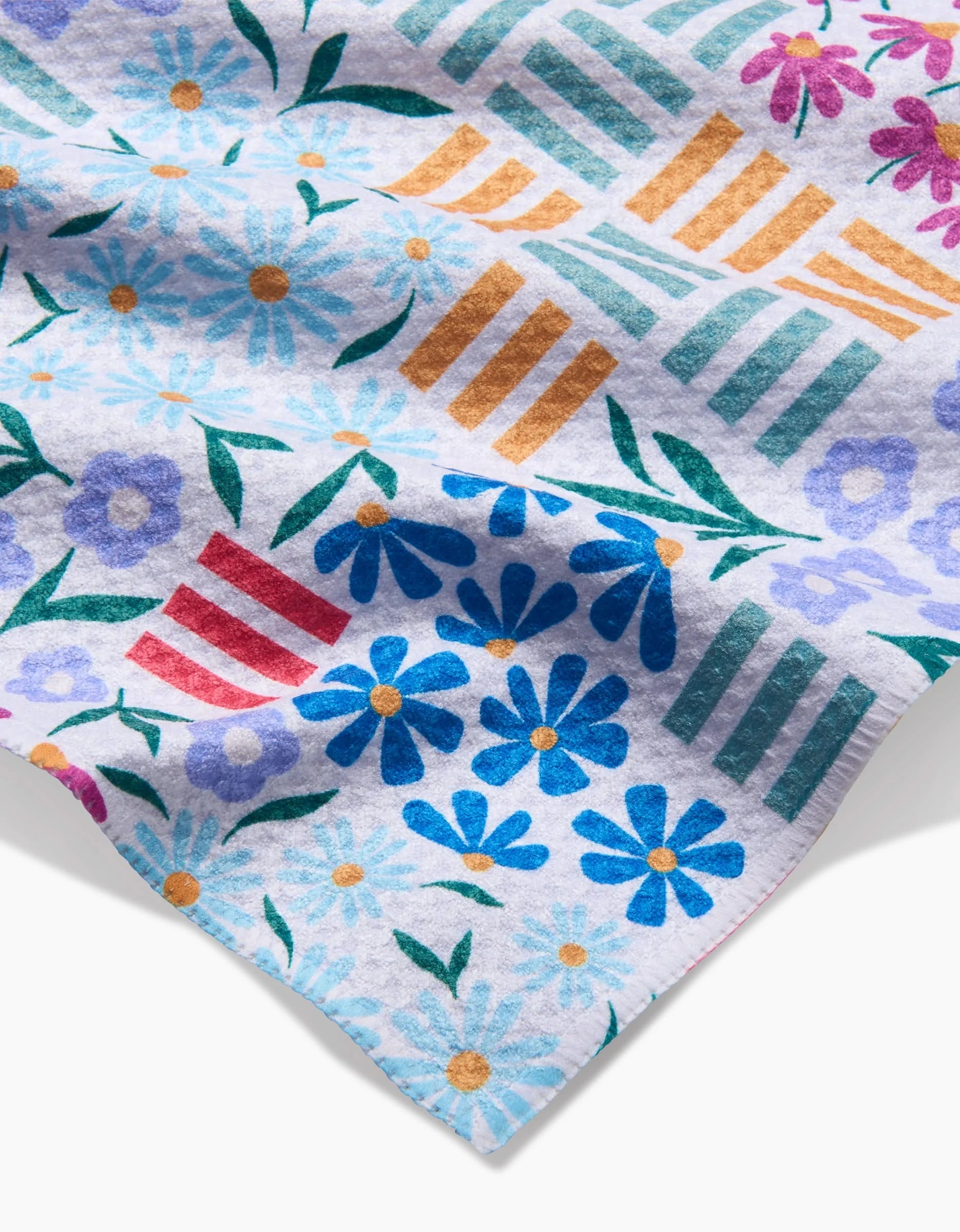 A close-up of Charlie Rowan Designs Floral Garden bar towel, featuring vibrant blue, red, and purple flowers, green leaves, and gold, teal, and red striped geometric shapes on a white Geoweave fabric background.