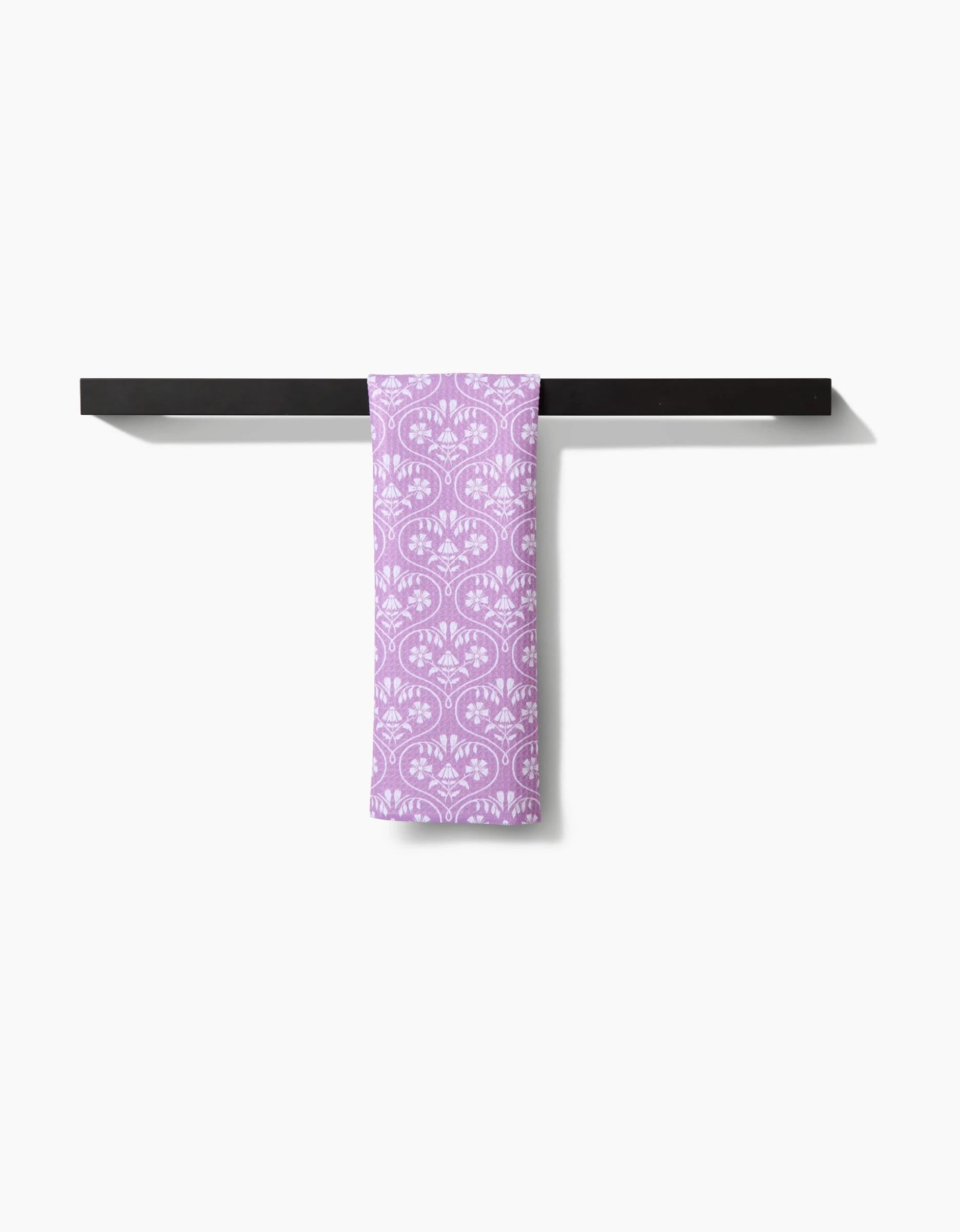 The “Floral Vine Hearts” bar towel by Hufton Studio, featuring a white floral pattern on lavender Geoweave fabric, hangs neatly over a black towel rack against a plain white background.