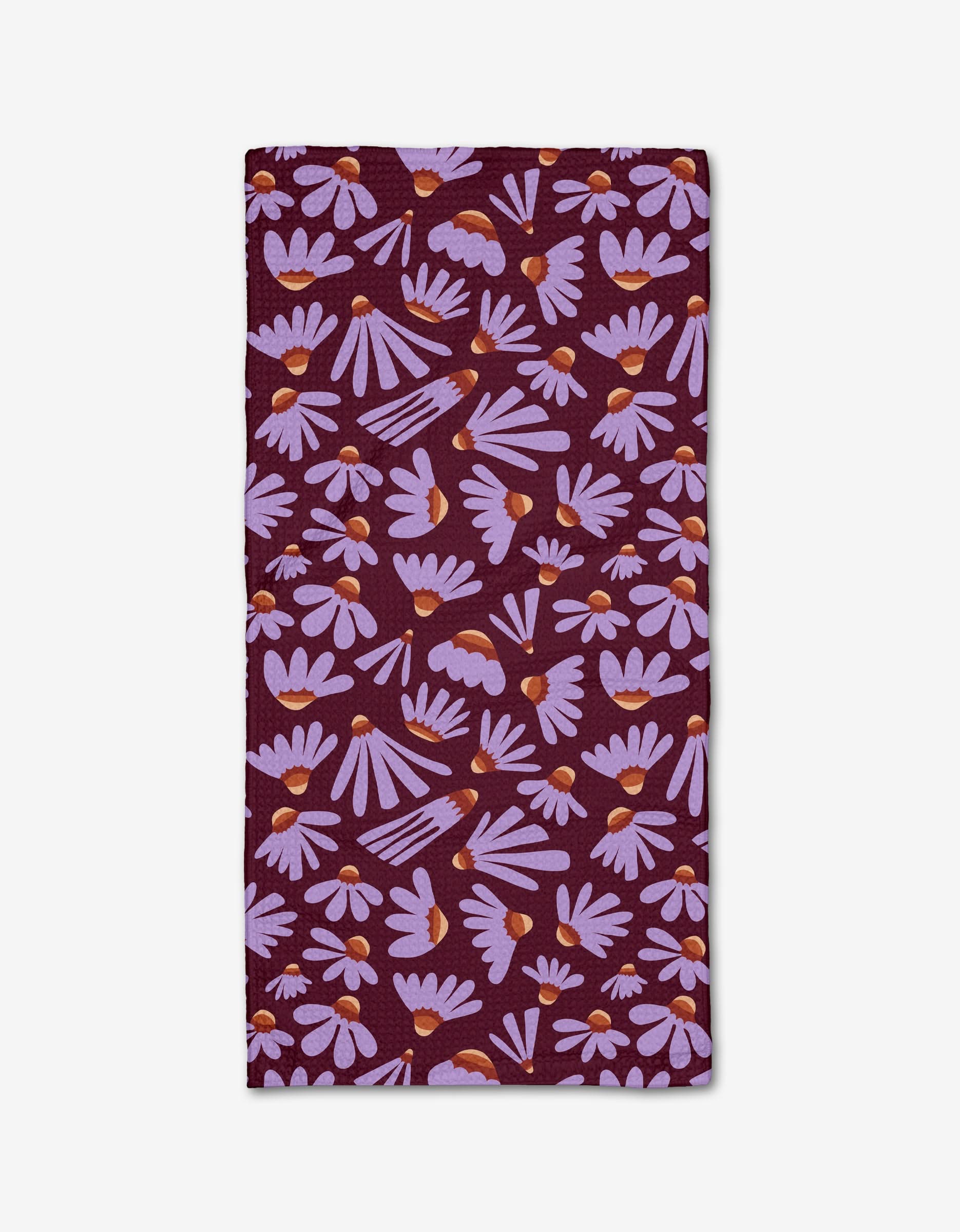 The Flower Pop napkin by Elisabeth Wing is a rectangular cloth made from recycled materials, featuring a dark purple background with light purple daisies and orange centers scattered across the surface.