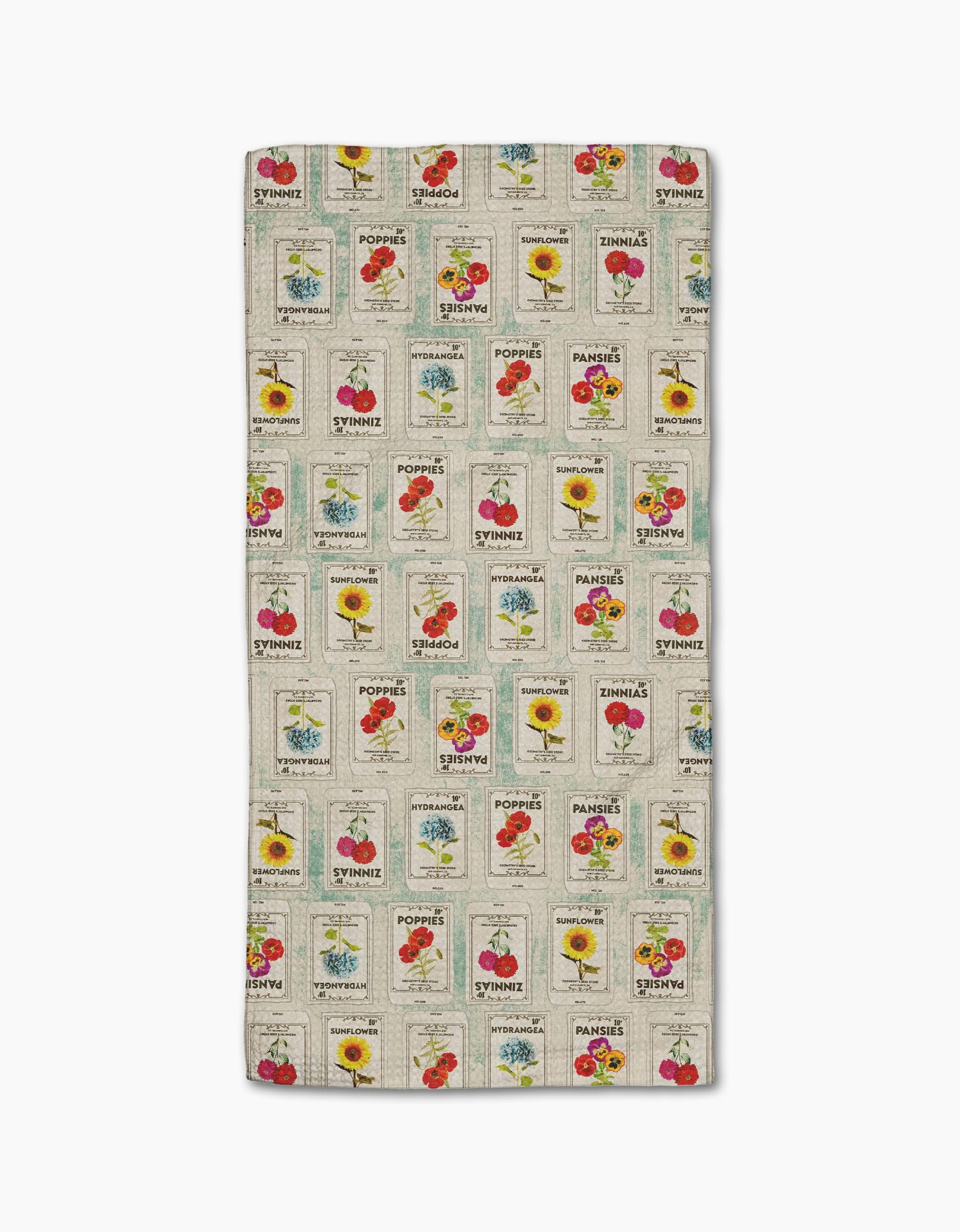 The Geometry Flower Seed Packs bar towel is rectangular, made from recycled materials, and features a vintage seed packet print with poppies, sunflowers, and tomatoes in a grid on a beige background.