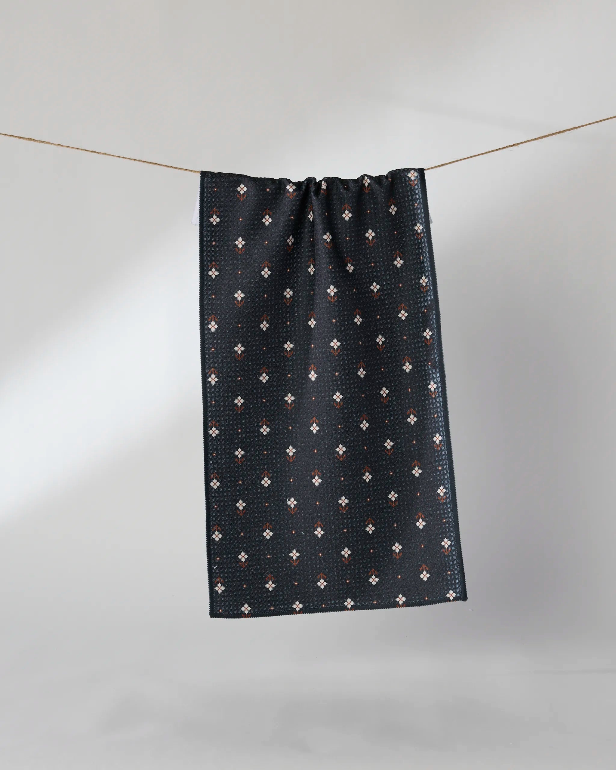 The Geometry Folk Pixel bar towel, featuring a small white and red floral pattern on a dark background, hangs on a clothesline against a plain, light backdrop.