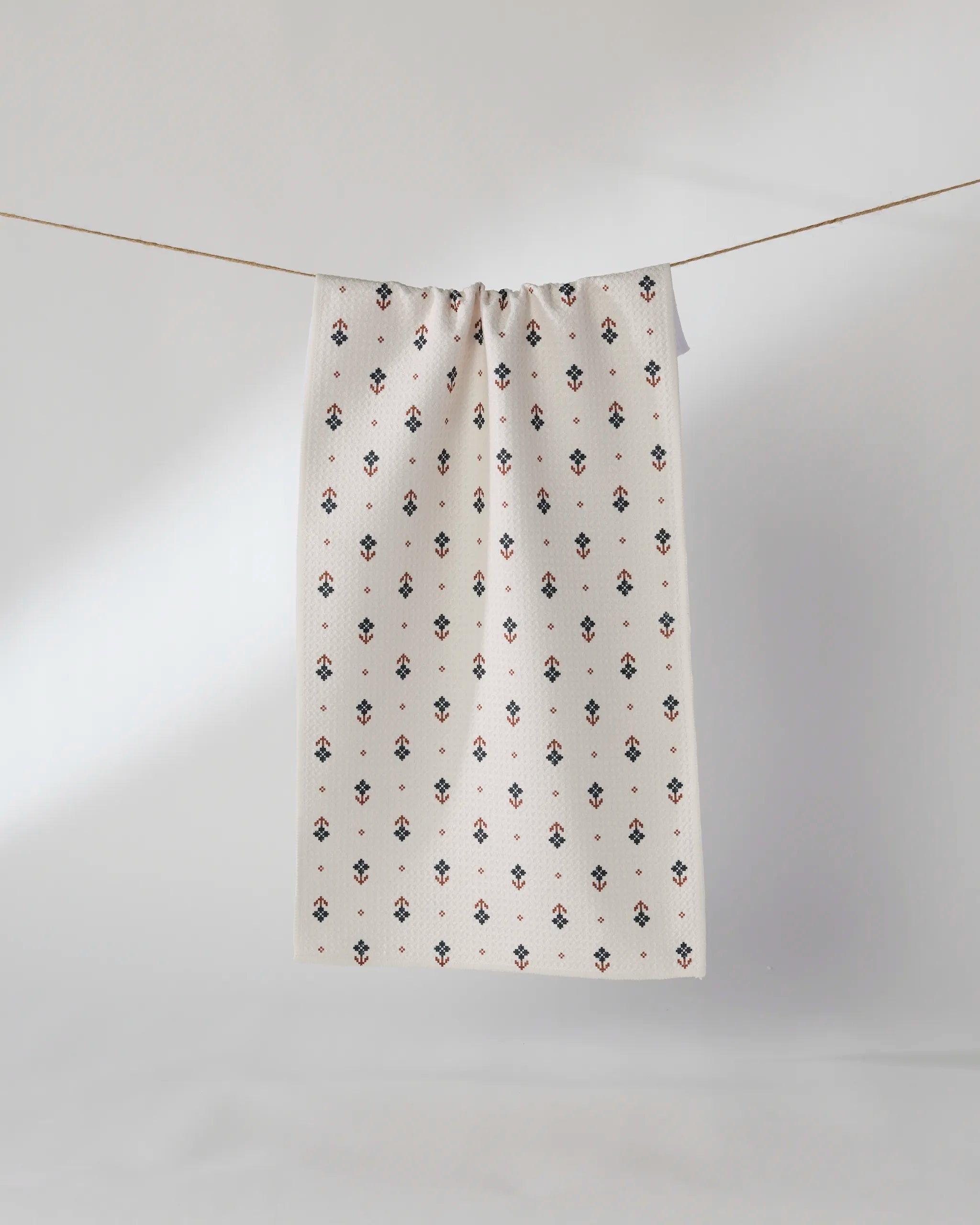 The Geometry Folk Pixel bar towel, featuring a white textured design with small black and red floral patterns, hangs on a thin clothesline against a minimal light background.