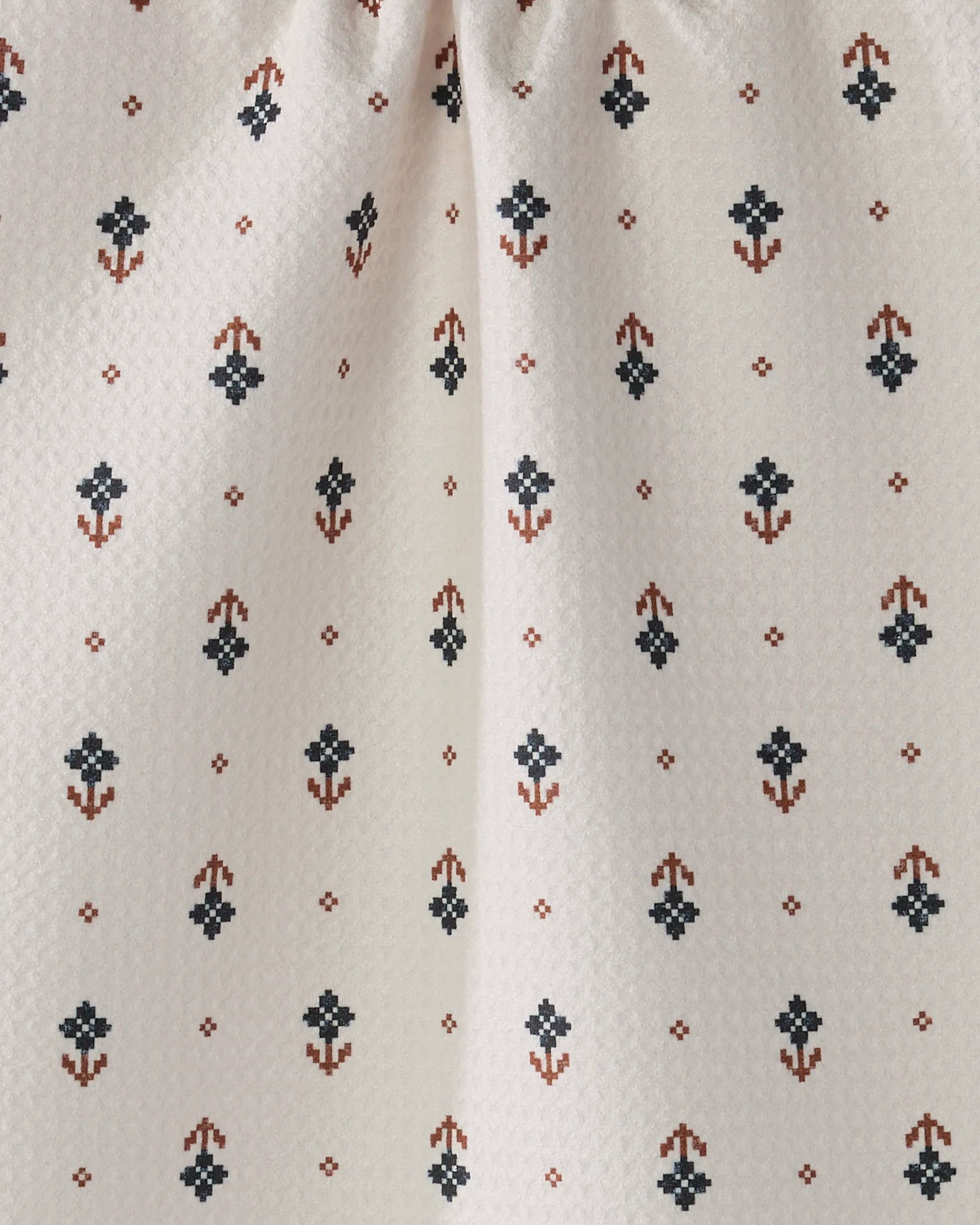 Close-up of Folk Pixel by Geometry: a cream-colored fabric made from recycled materials, featuring neat rows of small, stylized blue and brown floral motifs. The fabric appears slightly gathered near the top.