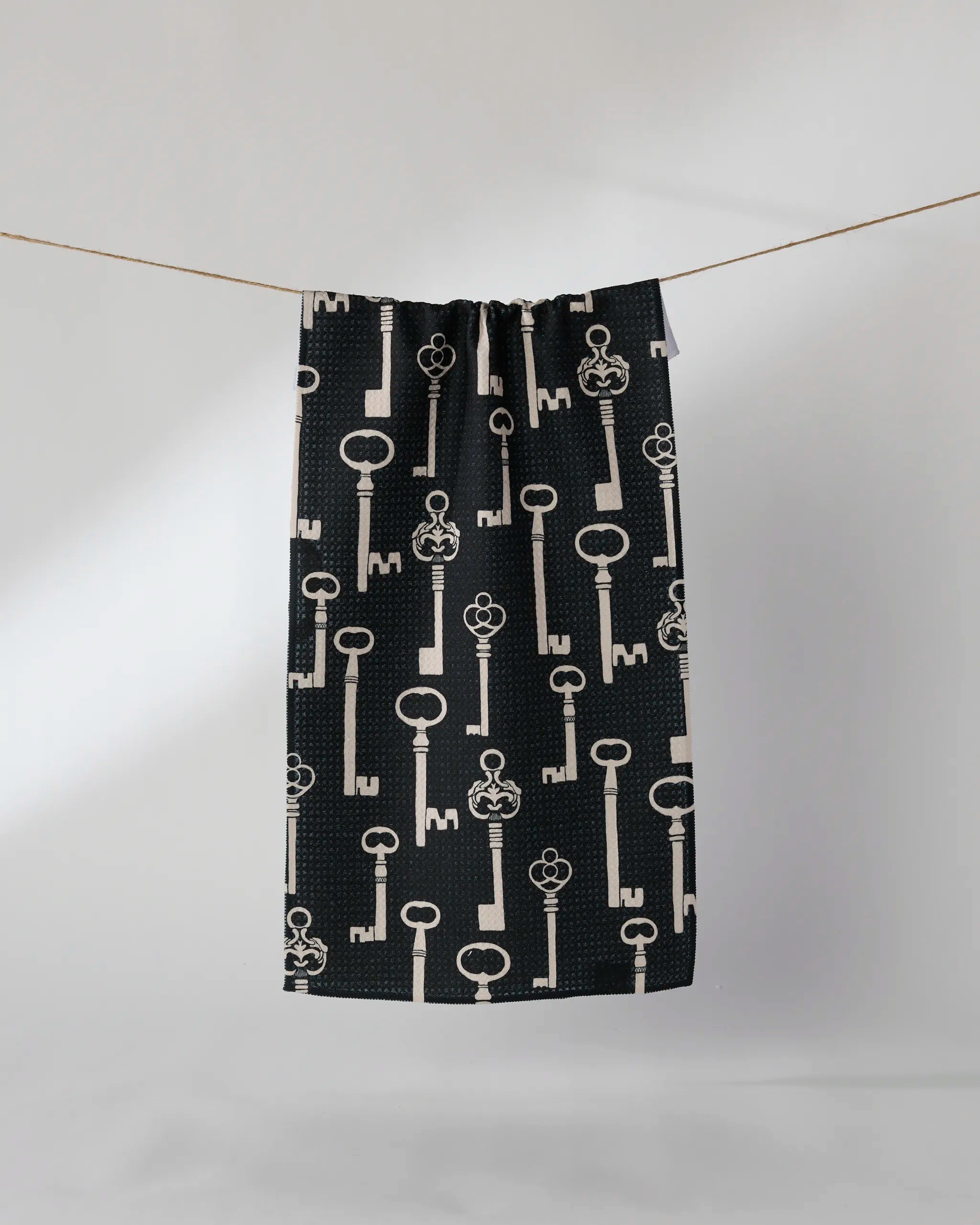 The Geometry "Forbidden Doors" towel, ultra-absorbent and black with a white vintage key pattern, hangs on a clothesline against a plain white background.