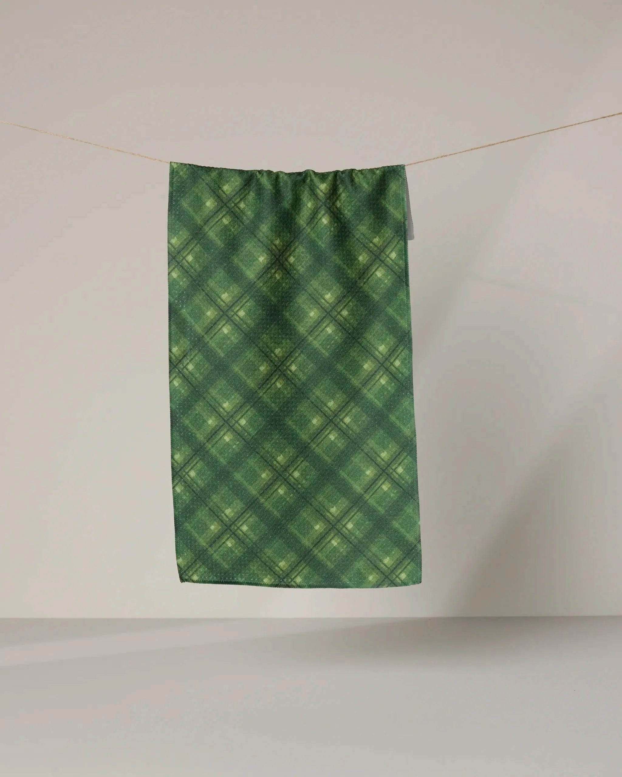 The Geometry Forever Plaid towel, featuring a green checkered pattern and ultra-absorbent design, hangs on a clothesline against a plain, light background.