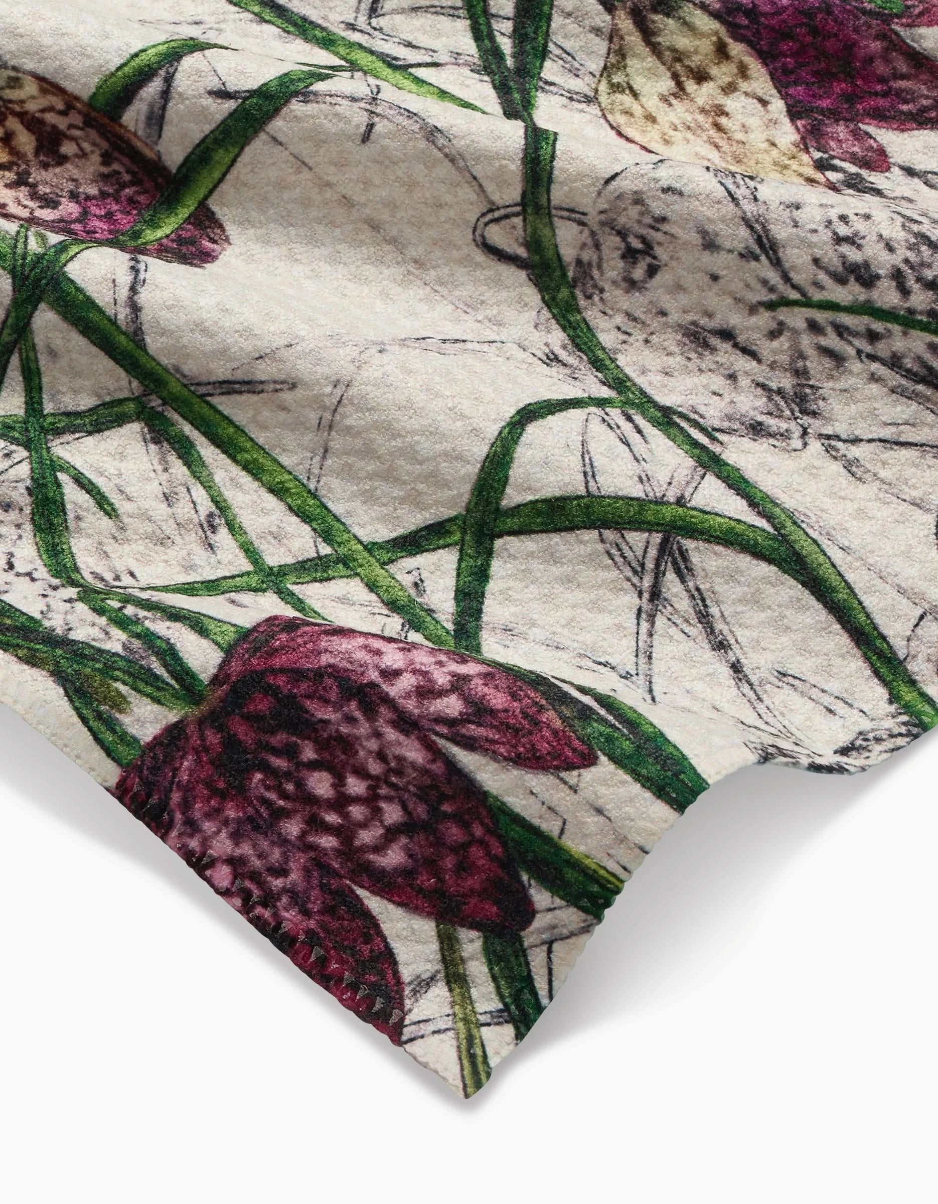 A close-up of Pascale Dilgers Fritillaria bar towel shows its Geoweave fabric with purple checkered flowers and green stems on a light background, featuring a folded edge that highlights its ultra-absorbent, soft texture.